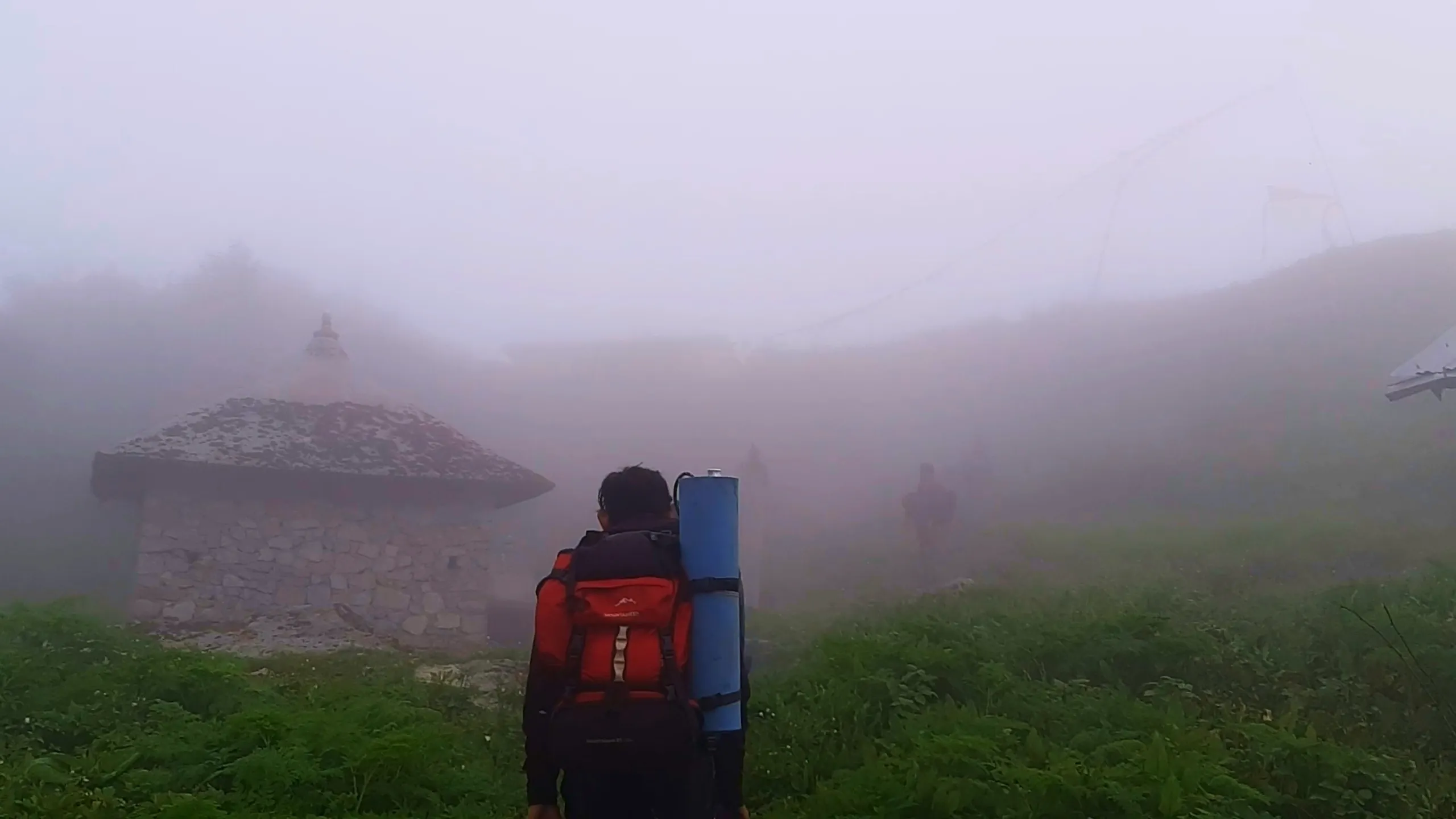 Atmospheric photo of a hiker walking away from the camera through thick white fog toward a traditional stone-built temple structure with a tiered pagoda-style roof, wearing a black jacket and carrying a red "Mountainer" backpack with a blue rolled sleeping mat. Lush green vegetation surrounds the path, with another stone structure and what appears to be a suspension bridge or cable structure faintly visible through the dense mist in the background, along with another distant figure. This captures the misty approach to the historic Narayanthan temple at Bethanchowk, Nepal Himalayan temple trekking, foggy mountain shrine visit, spiritual trekking destination Kathmandu hills.
