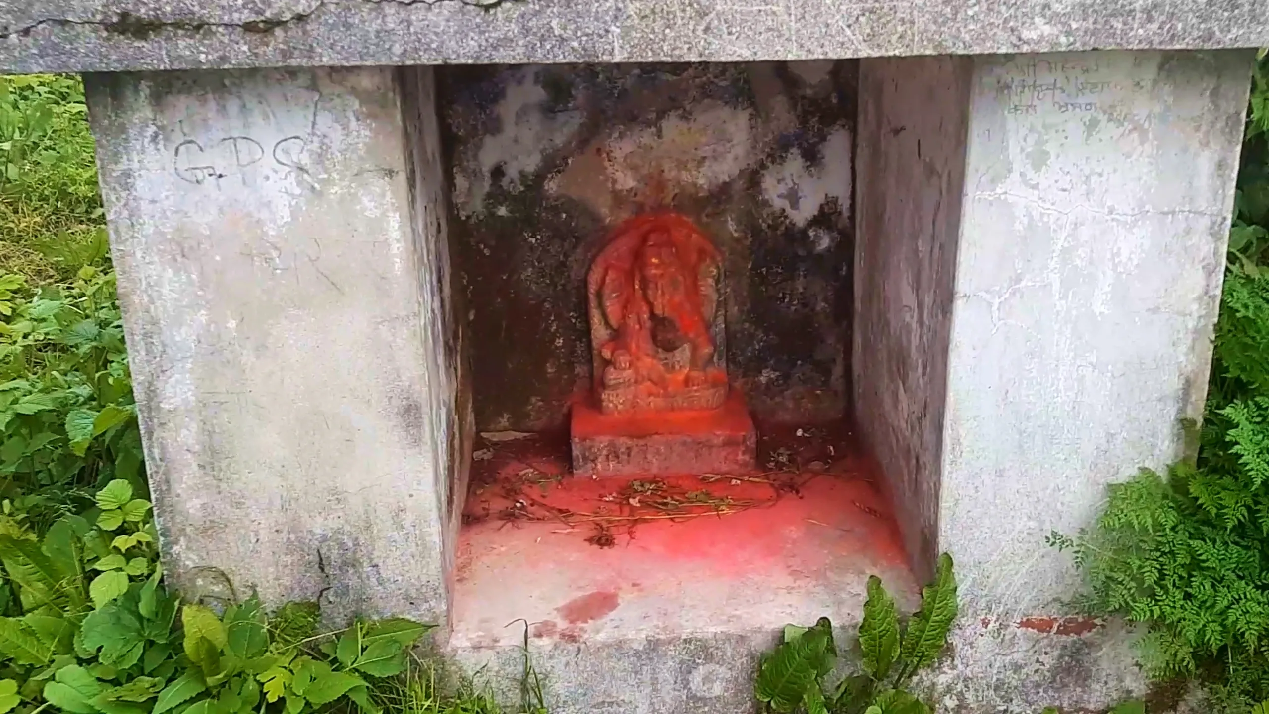 Close-up photo of a small weathered stone shrine niche carved into a concrete or stone structure, housing an ancient stone idol of a Hindu deity covered in bright red vermillion powder (sindoor), seated on a red-painted pedestal base scattered with dried flower offerings and twigs. The surrounding stone walls show dark moss and lichen staining with faded graffiti including the letters "GPS" visible on the left pillar. Green ferns and grass surround the shrine at ground level. This captures the sacred stone deity shrine at Narayanthan temple Bethanchowk, Nepal Hindu temple trekking destination, Himalayan religious site photography, spiritual pilgrimage trekking Kathmandu hills.