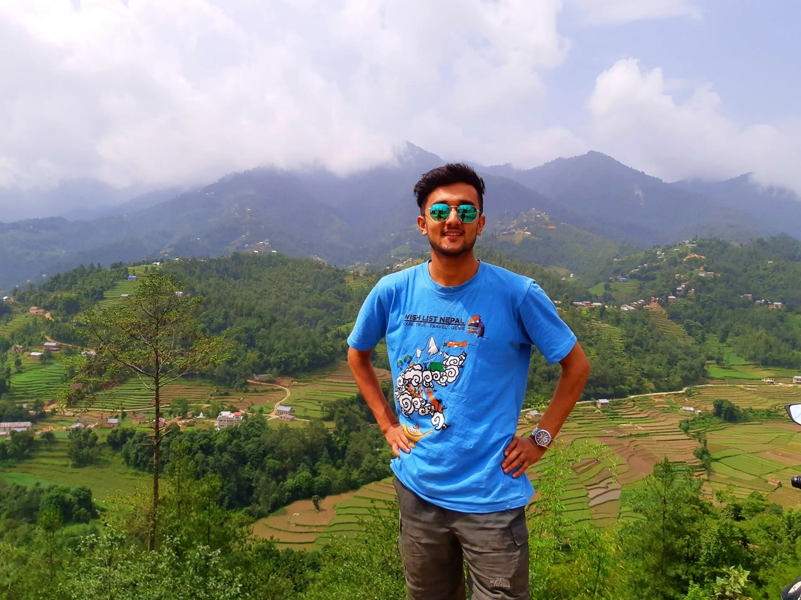 Portrait photo of a smiling young man wearing mirrored green and blue aviator sunglasses and a sky-blue "Wish List Nepal" t-shirt with a colorful Himalayan adventure graphic print, standing confidently with hands on hips on a hilltop viewpoint. Behind him stretches a breathtaking panoramic view of layered green terraced rice fields and farmland spreading across a valley, with scattered rural houses, dense forested hills, and dramatic low clouds wrapping around distant mountain peaks under a bright overcast sky. This image showcases stunning Bethanchowk viewpoint scenery, Kathmandu Valley terrace farming landscape, Nepal Himalayan adventure travel photography, best hiking spots near Kathmandu Nepal.