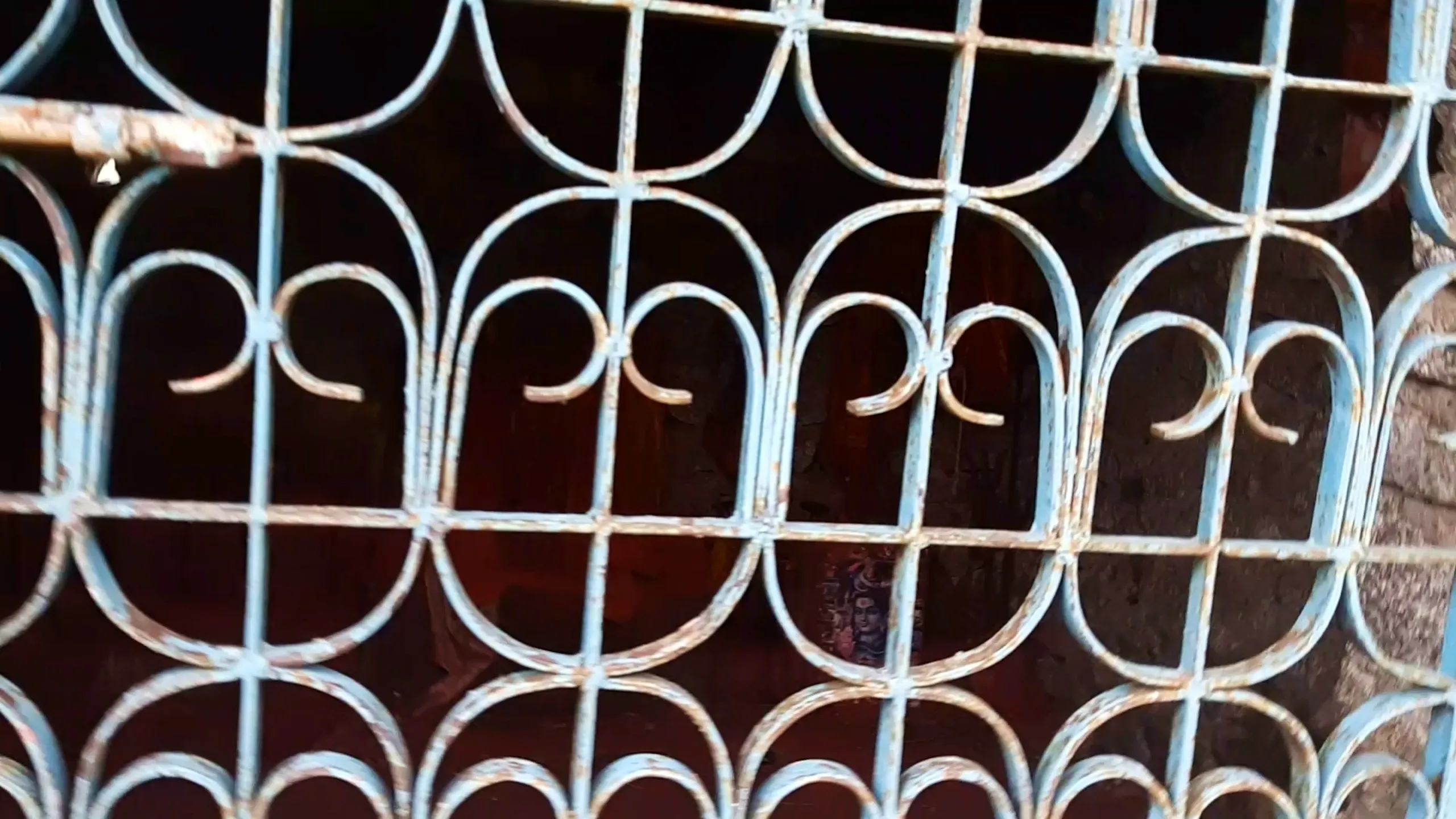 Extreme close-up photo of an ornate light blue painted decorative iron window grille with scrollwork and curved geometric patterns showing rust and peeling paint, mounted on a traditional building. Through the gaps in the metalwork, a dark interior space is visible with a small faded image or poster depicting a Hindu deity figure (possibly Lord Shiva) barely visible on the wall inside. This showcases the traditional decorative ironwork window at Narayanthan temple Bethanchowk, Nepal historic temple architecture, Himalayan religious building details, cultural heritage trekking site Kathmandu hills.