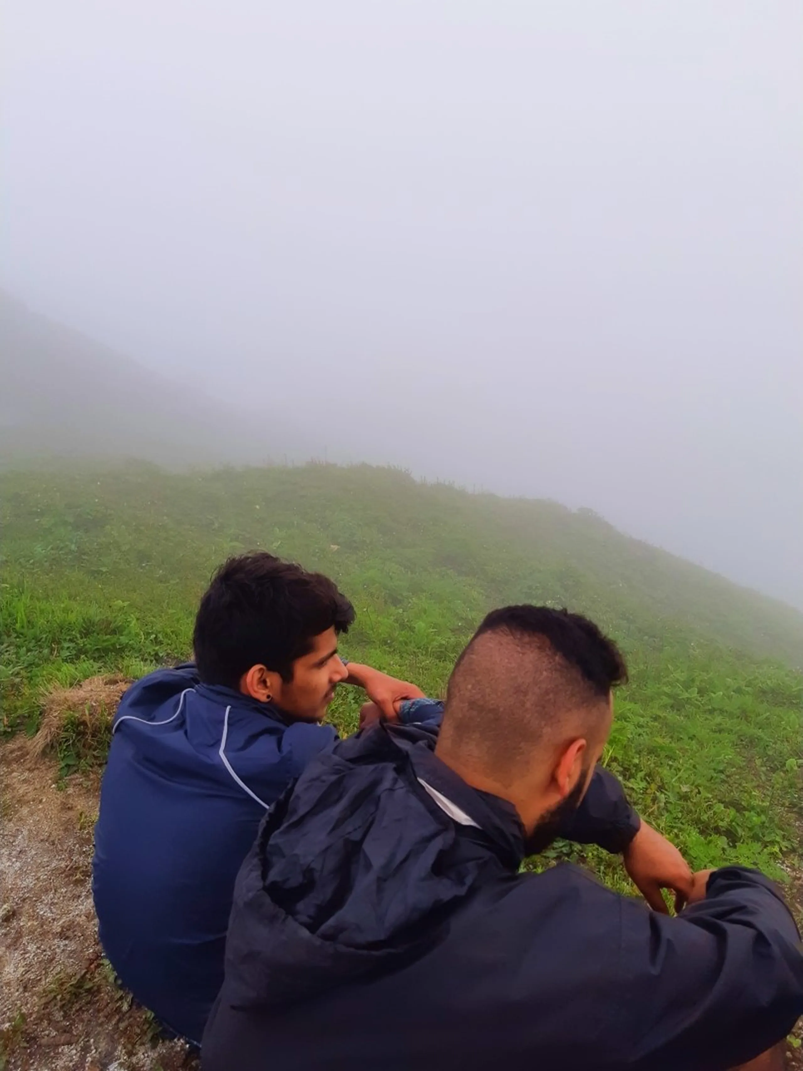 Quiet candid photo taken from behind of two friends sitting together on a misty grassy hillside ridge at Bethanchowk, gazing out into thick white fog covering the landscape below. The man on the left wears a navy blue jacket with white piping, while his friend in the foreground has a short undercut hairstyle with longer hair on top and wears a dark navy hooded jacket, resting his arm on his friend's shoulder. Green grass and ferns cover the slope beneath them, fading into complete whiteout fog. This captures a peaceful bonding moment overlooking the misty valley at Bethanchowk highest point, Nepal Himalayan foggy mountain views, trekking friends quiet moment, scenic overlook Kathmandu hills.