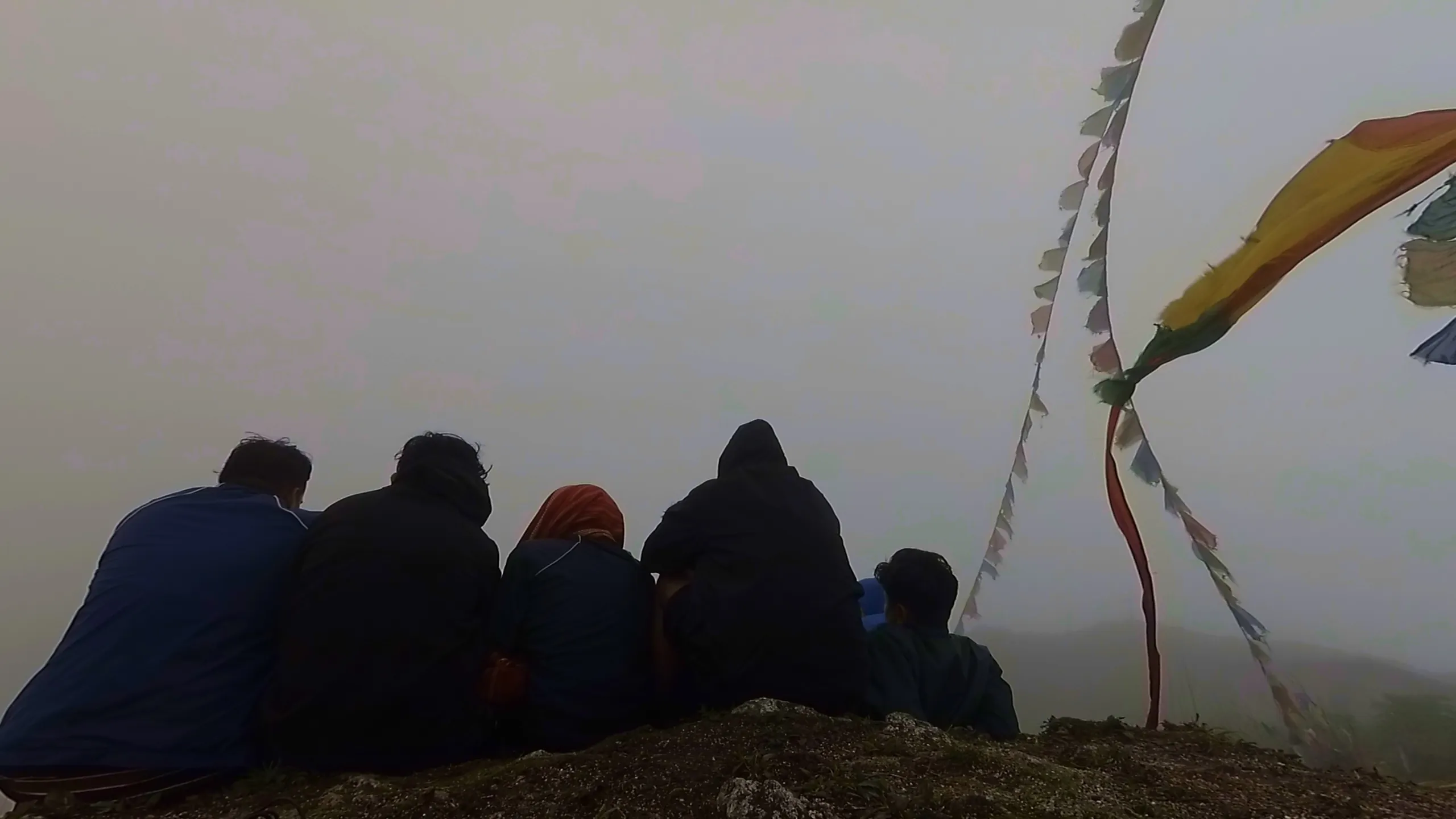 Silhouette photo of five young hikers sitting together in a row on a rocky hilltop edge at Bethanchowk, all facing away from the camera into thick white fog, wearing dark jackets including one in maroon and one in navy blue with a hood up. To the right, colorful Tibetan Buddhist prayer flags in red, yellow, blue, and white flutter dramatically from a tall wooden pole against the foggy white sky. This captures the spiritual summit moment with prayer flags at Bethanchowk highest point, Nepal Himalayan prayer flag photography, misty mountain peak trekking, group meditation viewpoint Kathmandu hills.