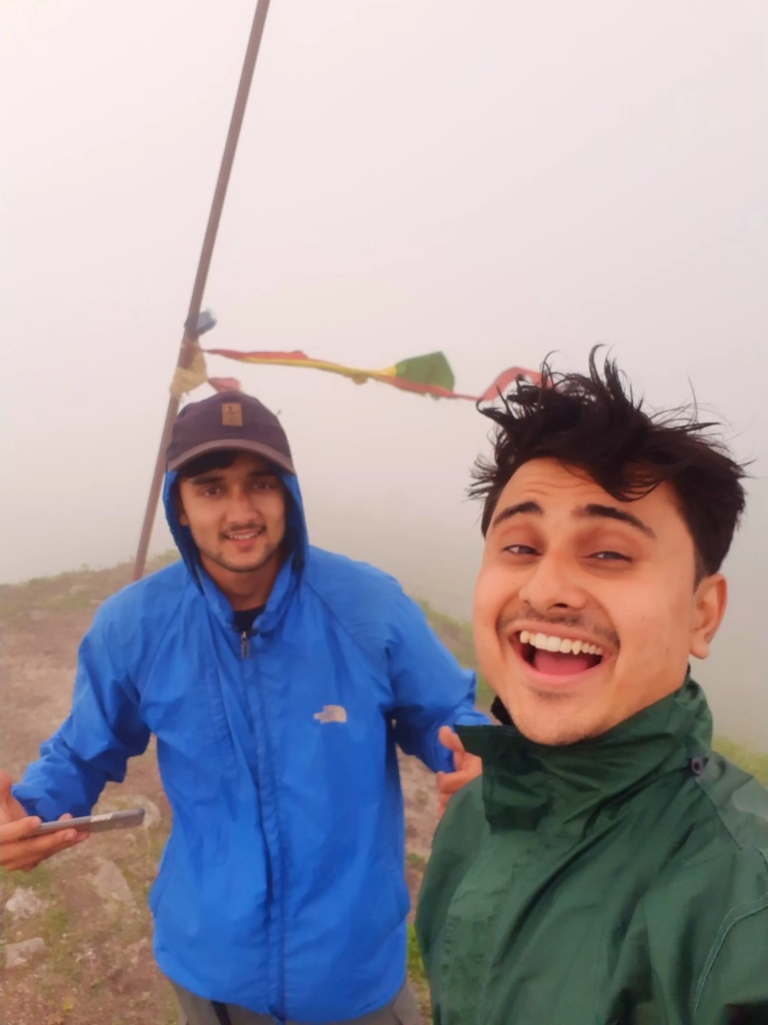 Cheerful close-up selfie of two young men at the foggy summit of Bethanchowk, Nepal, near a tall prayer flag pole with colorful red, yellow, and green Buddhist prayer flags fluttering behind them. The man on the right in front has wildly tousled dark hair and wears a green hooded rain jacket, laughing with mouth wide open. Behind him, another friend wearing a navy blue cap and matching blue "The North Face" rain jacket smiles gently, holding a smartphone. Thick white fog completely covers the background. This captures a joyful summit selfie at Bethanchowk prayer flag pole, Nepal Himalayan trekking celebration, misty mountain peak photography, group adventure memories Kathmandu hills.