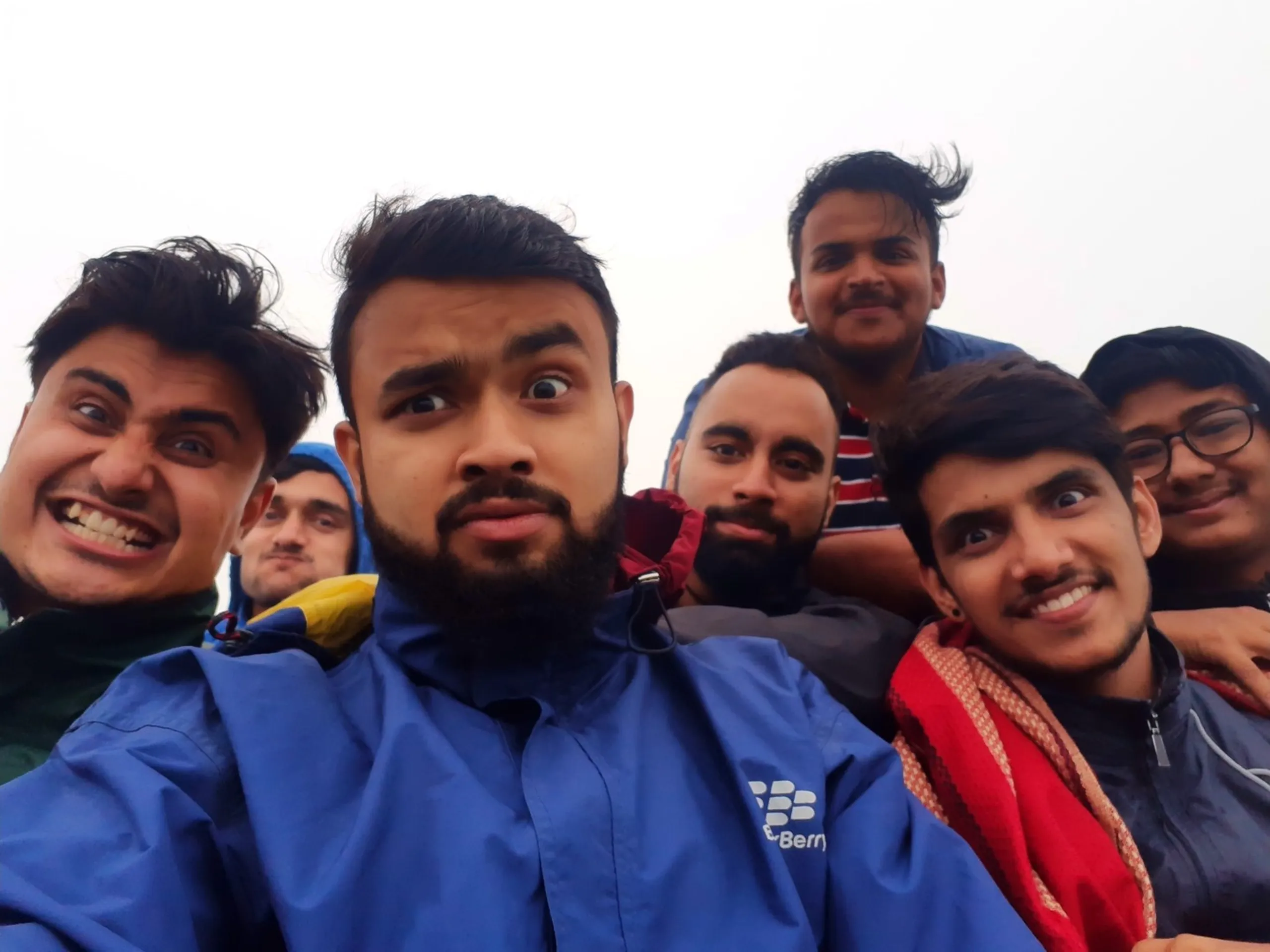 Large group selfie photo of seven young male friends huddled together at the foggy summit of Bethanchowk, Nepal, all making playful exaggerated facial expressions for the camera. The man in front wears a bright blue "BlackBerry" branded rain jacket with a wide-eyed surprised expression and full beard, surrounded by friends wearing green, navy, and grey jackets, one with a red scarf draped around his neck and another wearing glasses. Completely white foggy sky fills the background with no visible landscape. This captures a hilarious group summit photo at Bethanchowk highest point, Nepal trekking friends funny moments, Himalayan misty mountain group selfie, adventure trip memories Kathmandu hills.