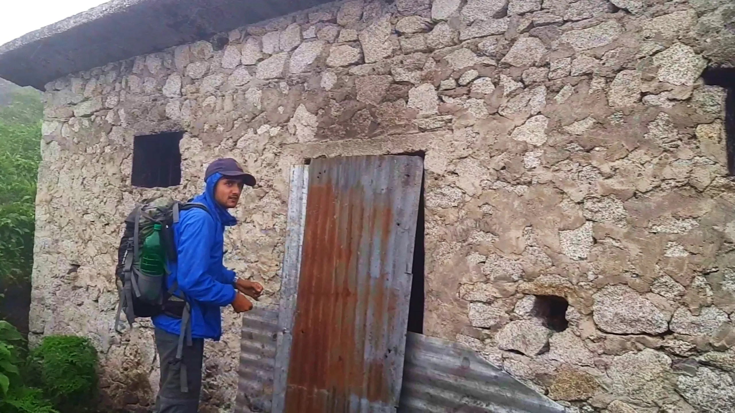 Photo of a young man wearing a blue rain jacket with hood up, a navy cap, and a hiking backpack with a green water bottle attached, standing beside the rusted corrugated metal door of an old stone cottage at Bethanchowk, looking back at the camera with a curious expression. The rubble-stone wall of the building shows weathered grey and tan stones with dark window openings. Green forest vegetation is visible on the left edge of the frame. This captures exploration of the abandoned stone shelter at Bethanchowk highest point, Nepal Himalayan mountain hut investigation, historic stone building trekking, adventure exploration Kathmandu hills.