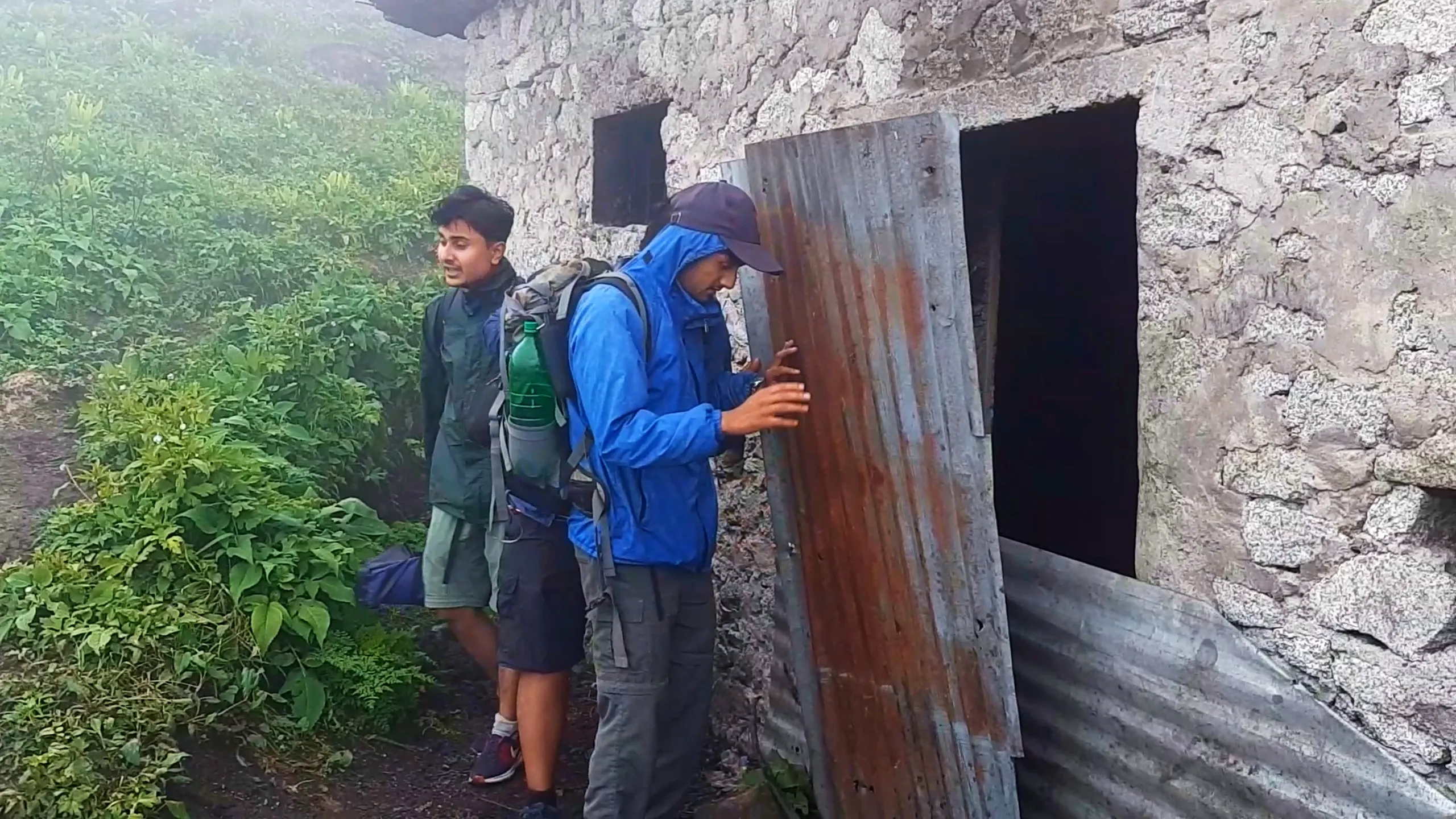 Photo of two young hikers standing beside the rusted corrugated metal door of an old stone cottage at Bethanchowk, with one man in a blue rain jacket, navy cap, and grey backpack reaching toward the door while another friend in a dark green jacket and grey cargo shorts with red sneakers stands behind him looking on. The weathered rubble-stone wall with dark window openings forms the backdrop, with green forest vegetation visible on the left. This shows friends investigating the historic stone shelter building at Bethanchowk highest point, Nepal Himalayan mountain hut exploration, abandoned cottage trekking landmark, group adventure discovery Kathmandu hills.