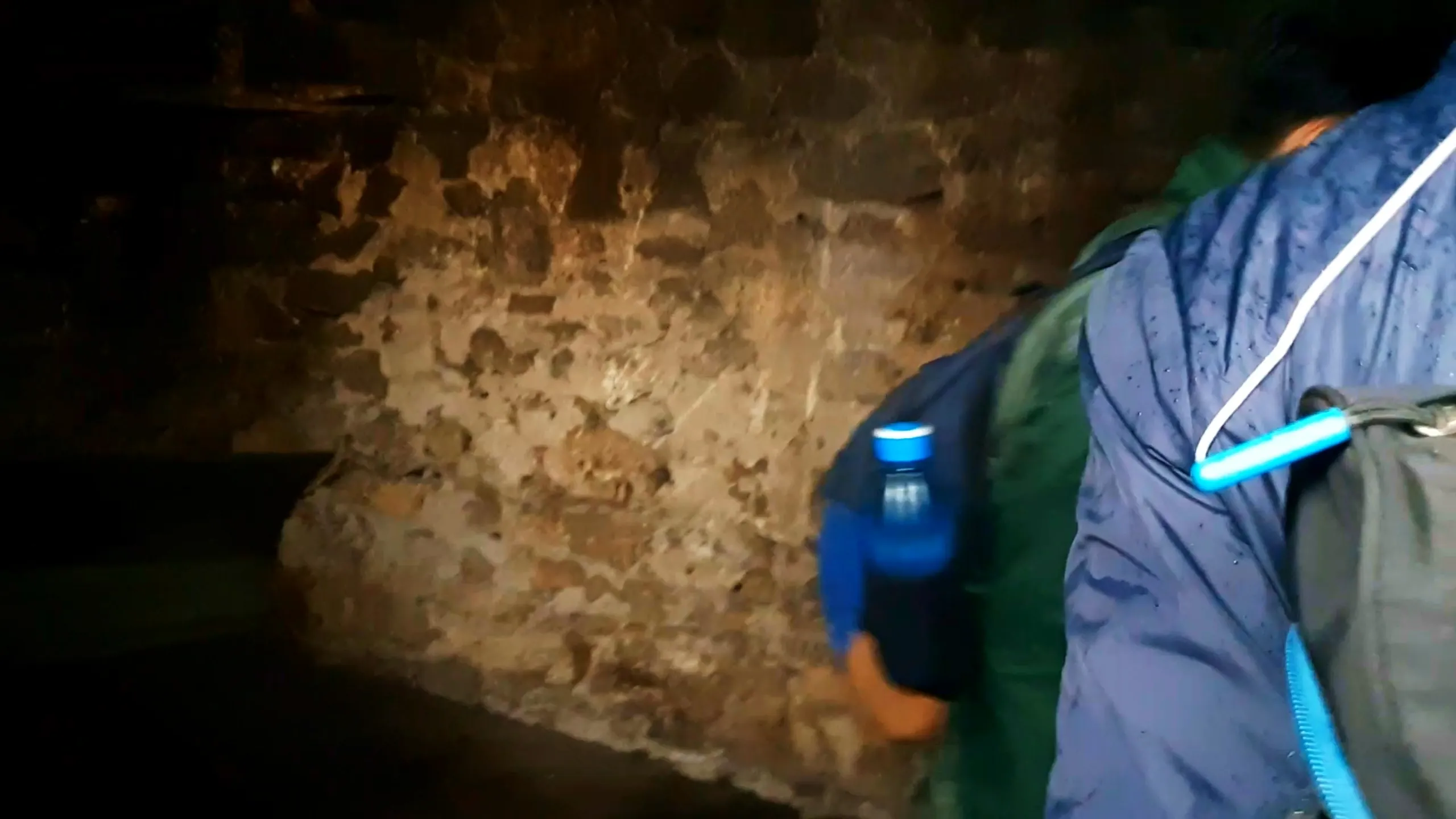 Dark blurry photo taken inside the old stone cottage at Bethanchowk showing the rough textured interior stone walls illuminated by a flashlight or camera light, with two hikers wearing blue and green rain jackets and backpacks with a blue water bottle attached, moving through the dim interior space. The weathered stone construction shows centuries-old craftsmanship with visible mortar and uneven rock surfaces. This captures the mysterious dark interior of the historic stone shelter at Bethanchowk highest point, Nepal Himalayan mountain hut interior, abandoned building exploration trekking, adventure discovery Kathmandu hills.