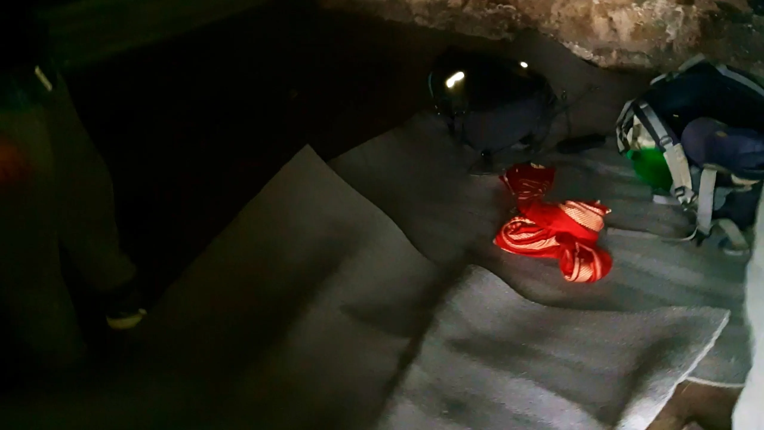 Dark photo of the interior floor of the old stone cottage at Bethanchowk, showing a grey foam sleeping mat laid out on the ground with a black motorcycle helmet, a red bundled jacket or bag, and a backpack with a green water bottle attached resting nearby. The rough stone wall and ceiling are visible in dim lighting at the top of the frame. This shows the makeshift overnight camping setup inside the historic stone shelter at Bethanchowk highest point, Nepal Himalayan mountain hut camping, indoor shelter trekking accommodation, adventure overnight stay Kathmandu hills.