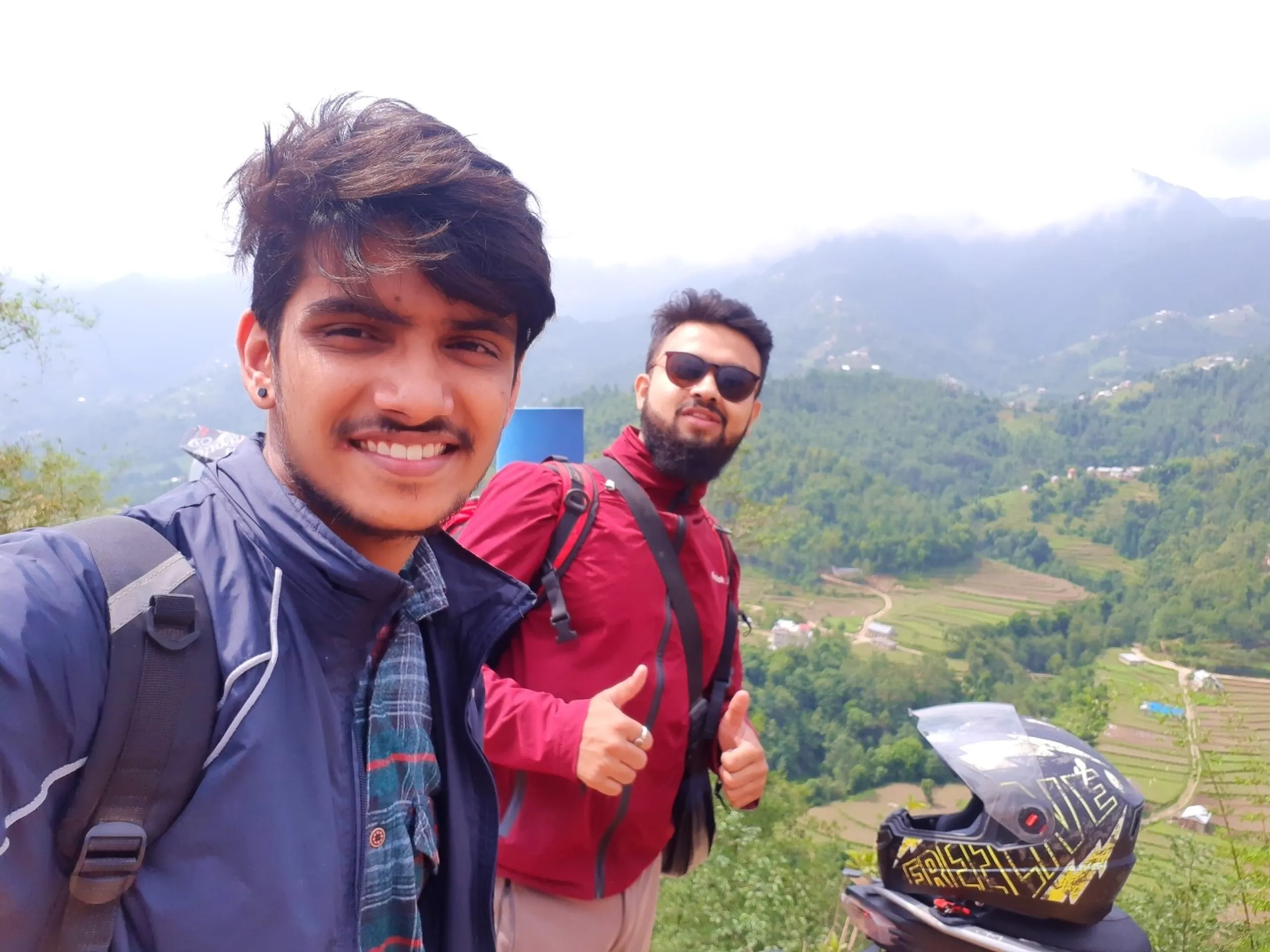 Close-up selfie of two young male friends smiling at a misty mountain viewpoint during a trekking trip in Nepal. The man in front has tousled brown hair, an ear piercing, and wears a navy blue rain jacket with a plaid shirt underneath and a grey backpack strap visible. Behind him, a bearded friend wearing black sunglasses and a maroon red rain jacket gives two thumbs up gesture. In the foreground, a black and yellow graphic motorcycle helmet rests on a bike seat. The backdrop shows expansive green terraced hillside farmland fading into thick white fog and clouds covering distant mountains. This captures a joyful bonding moment during a Bethanchowk hiking trip, friends trekking Nepal, Himalayan misty mountain views, motorbike trekking adventure Kathmandu.