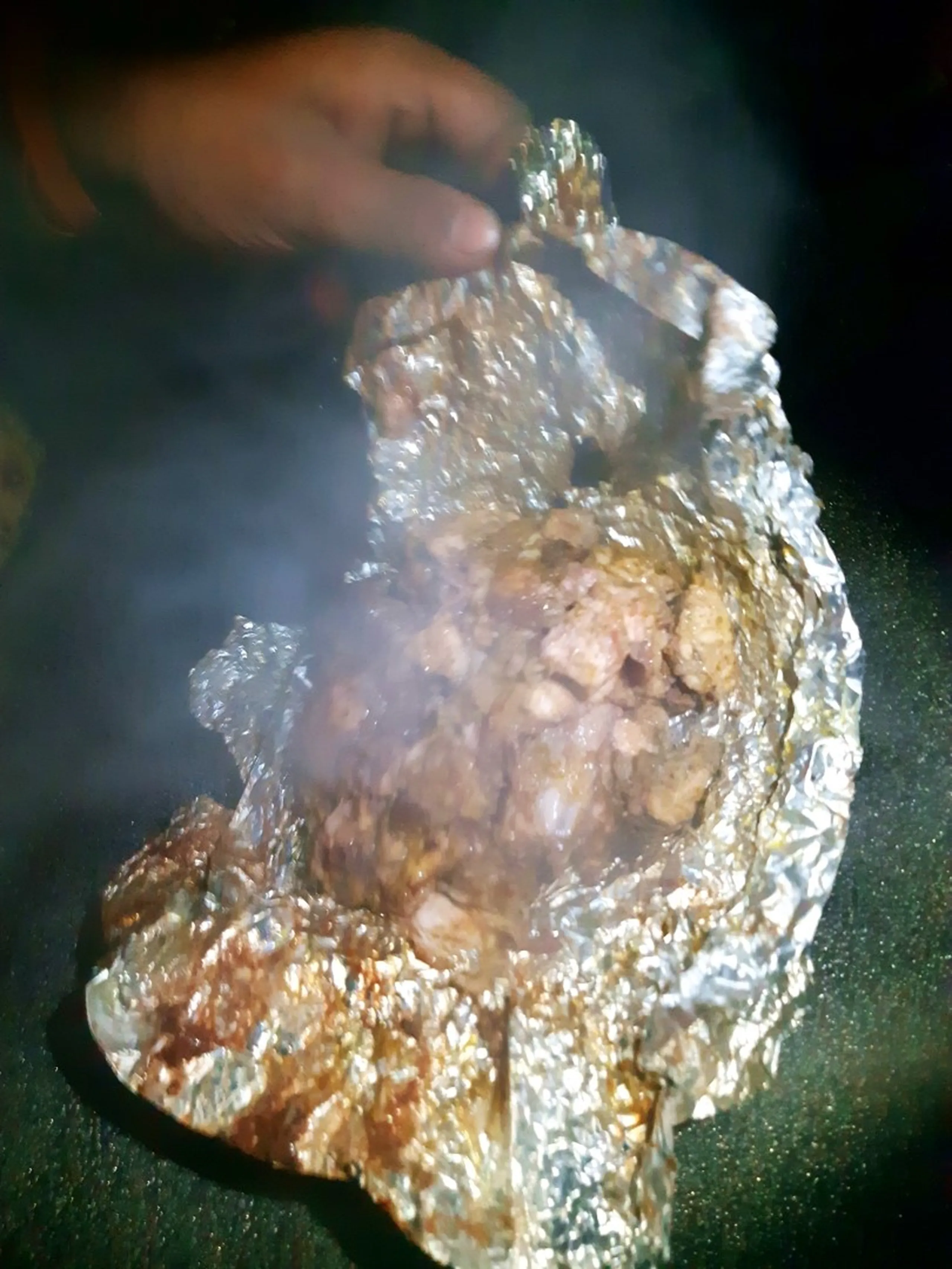 Close-up nighttime photo of a hand holding open a piece of aluminum foil containing roasted or grilled meat pieces with golden-brown caramelized edges, steam rising from the hot food, photographed in dim firelight with a dark smoky background. This showcases the campfire-cooked meal preparation during the Bethanchowk overnight camping trip, Nepal jungle camping food, Himalayan trekking BBQ meal, outdoor cooking adventure Kathmandu hills.