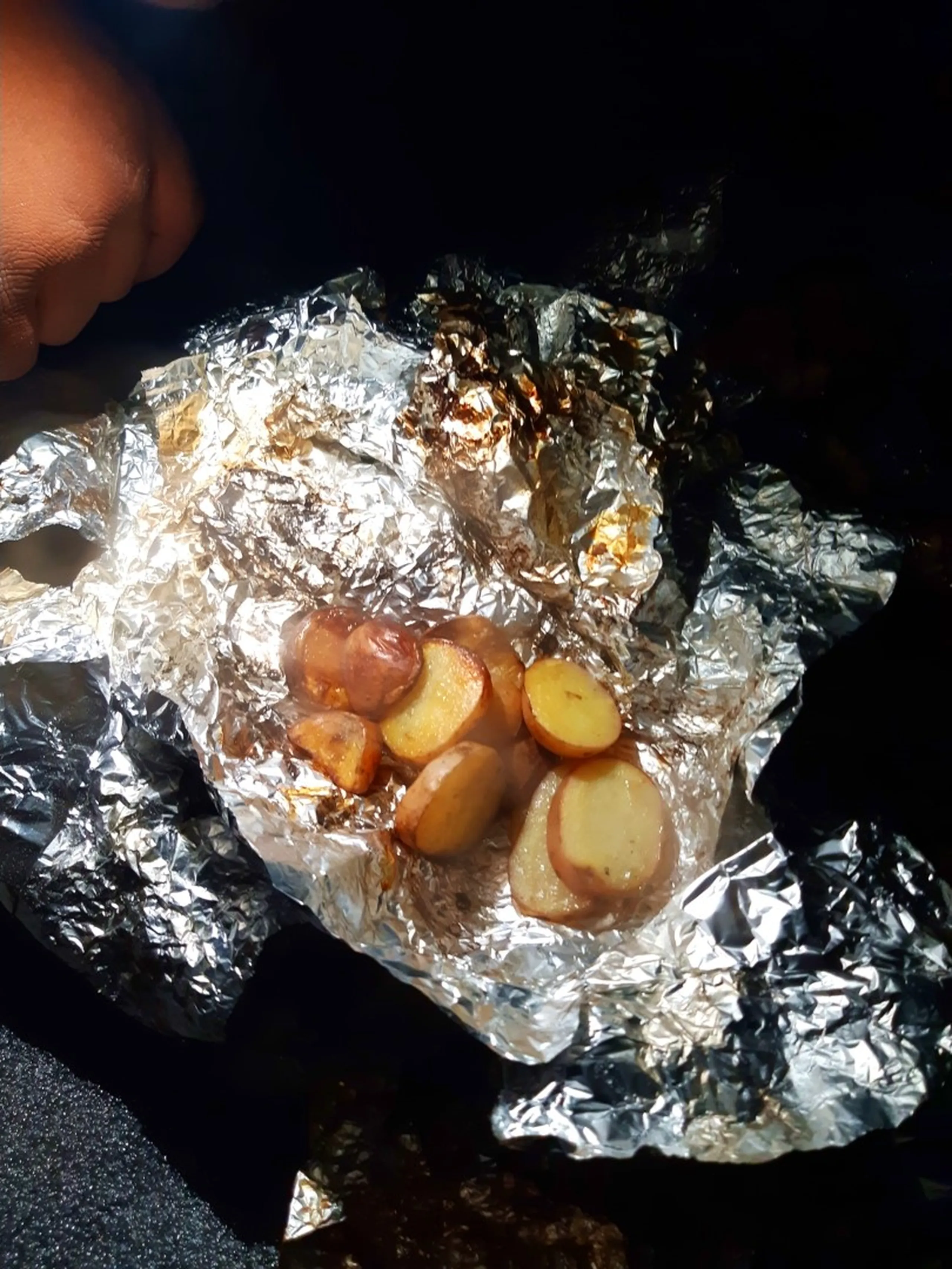 Close-up nighttime photo of aluminum foil packet opened to reveal roasted potato wedges with crispy golden-brown and red skin, cooked over an open campfire, held by a hand in the dim firelit darkness. This captures the rustic campfire-roasted potato dish during Bethanchowk overnight camping trip, Nepal jungle camping recipes, Himalayan trekking food preparation, outdoor cooking adventure Kathmandu hills.