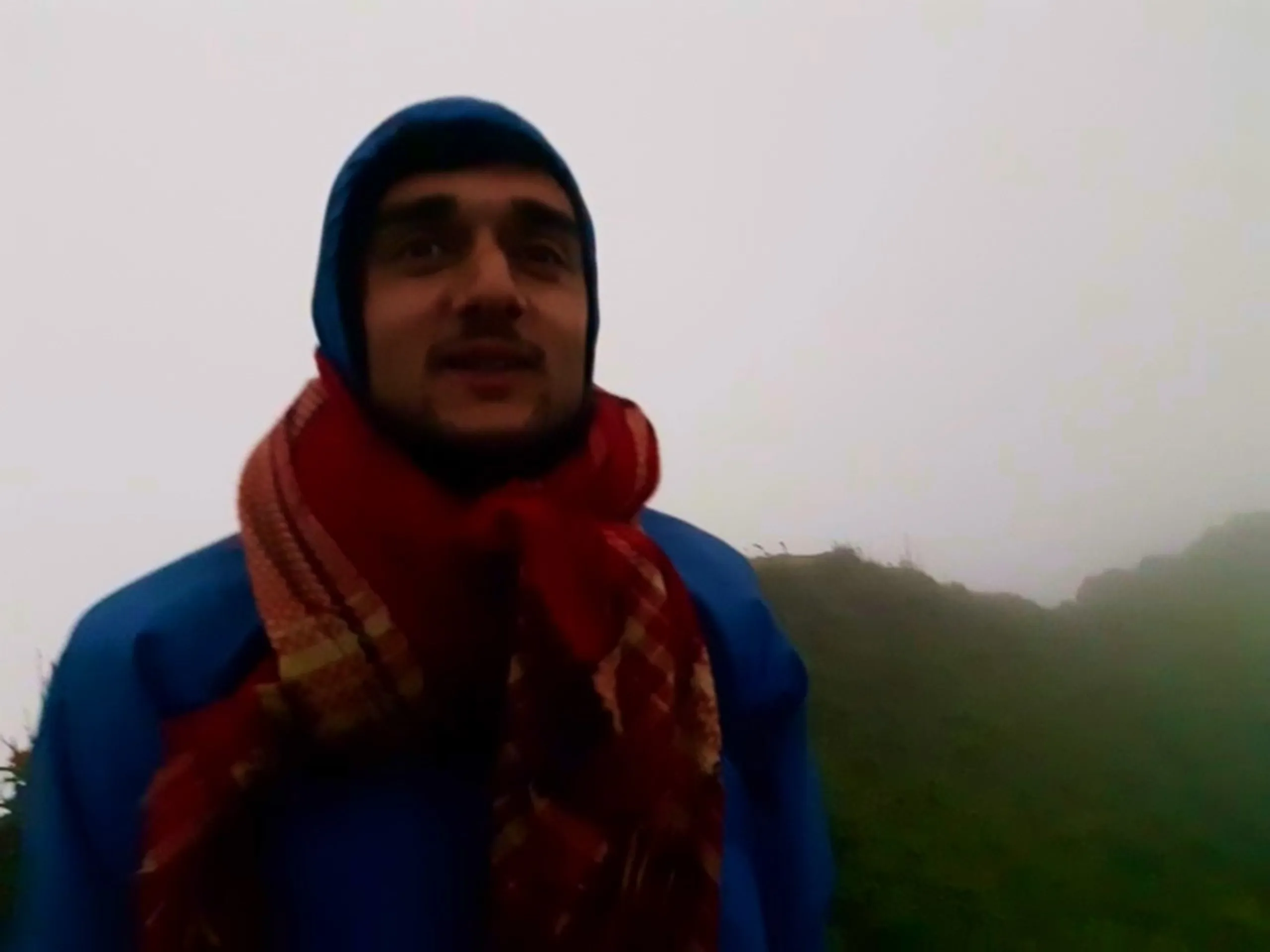 Close-up portrait of a young man wearing a blue hooded rain jacket with a red and orange plaid scarf wrapped around his neck and lower face, looking at the camera with a slight smile while talking, against a backdrop of thick white fog and a glimpse of green grassy hillside on the right. This captures a cold misty morning portrait at Bethanchowk highest point, Nepal Himalayan foggy mountain trekking, cold weather hiking gear, adventure travel portrait Kathmandu hills.