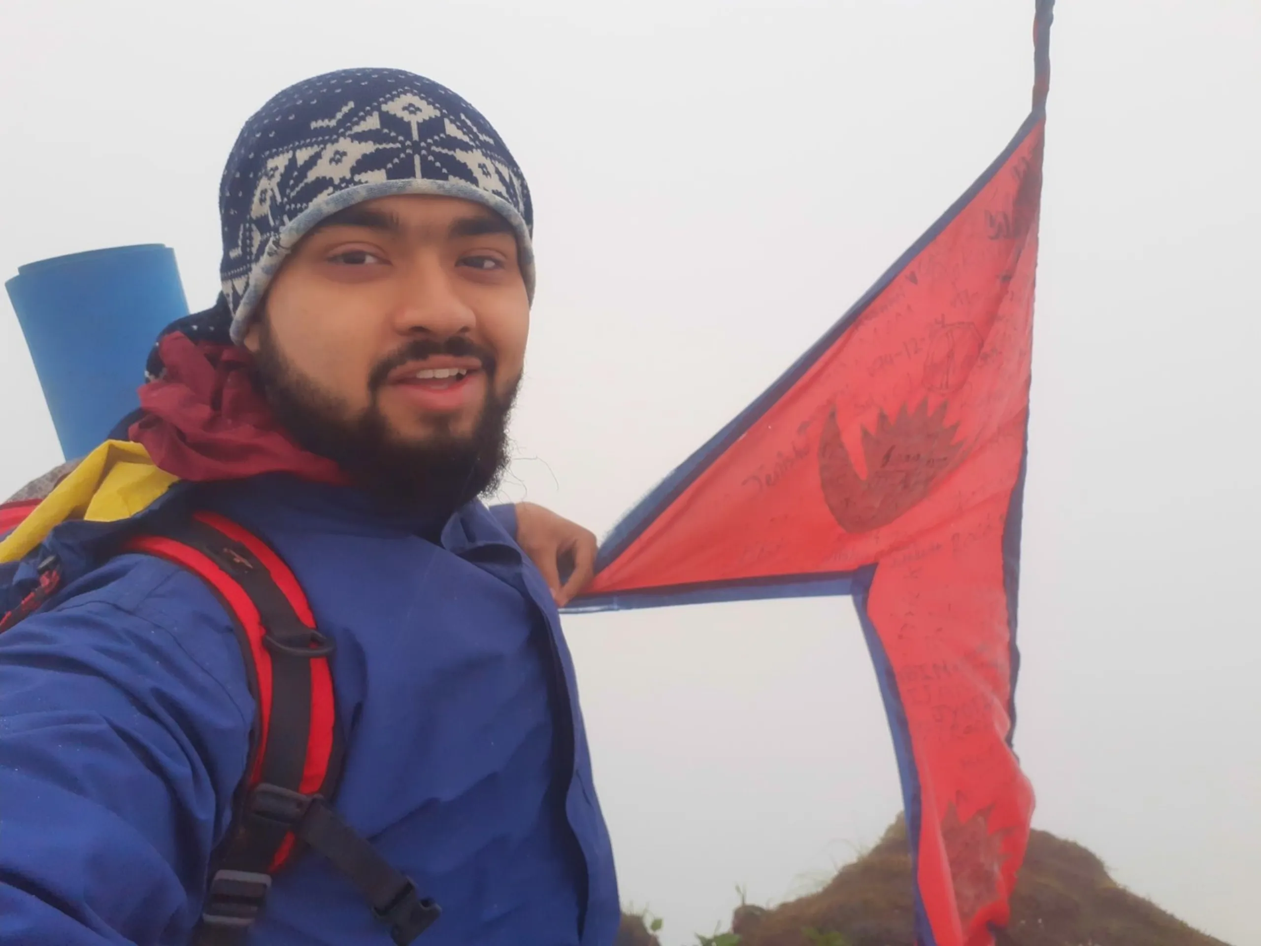 Close-up selfie of a bearded young man wearing a navy and white patterned knit beanie hat and a blue jacket with a maroon and yellow garment draped over his shoulder, smiling and posing beside a large red Nepali national flag with the distinctive double-pennant crescent moon and sun symbols, covered in handwritten signatures and messages from previous trekkers. A blue rolled sleeping mat is visible on the left edge. Thick white fog fills the background. This captures the iconic Nepali flag summit photo at Bethanchowk highest point, Nepal flag trekking achievement, Himalayan summit celebration, mountain peak photography Kathmandu hills.