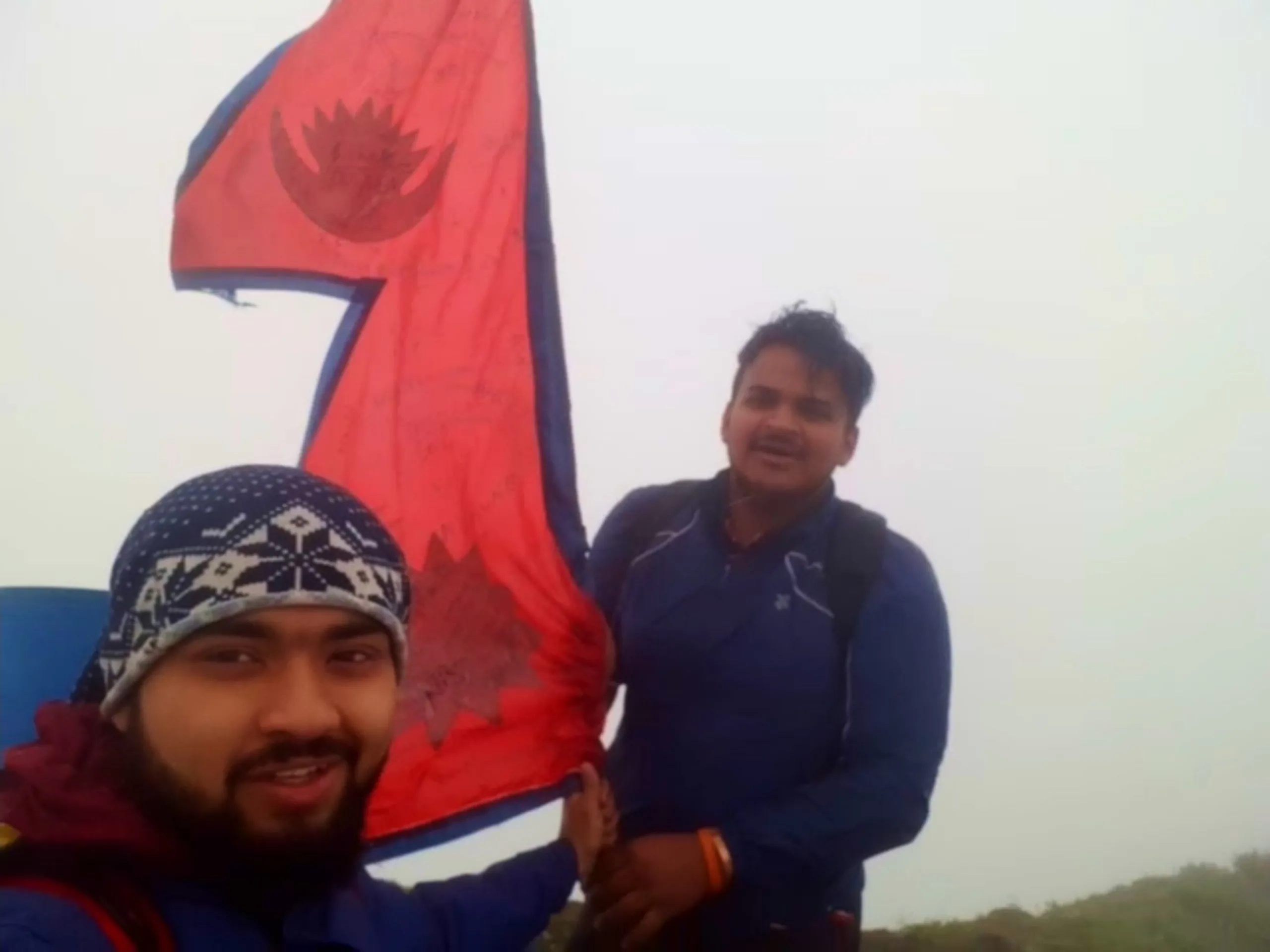 Photo of two young men posing beside a large red Nepali national flag with the crescent moon and sun emblem, covered in handwritten signatures from past trekkers, fluttering against a completely white foggy sky. The man in front wears a navy and white patterned beanie and blue jacket, smiling at the camera, while his friend behind wears a navy blue zip-up jacket with a backpack, looking slightly upward with a content expression. This captures the signature-covered Nepal flag landmark at Bethanchowk highest point, Nepal Himalayan trekking flag photo, mountain summit tradition, adventure travel milestone Kathmandu hills.