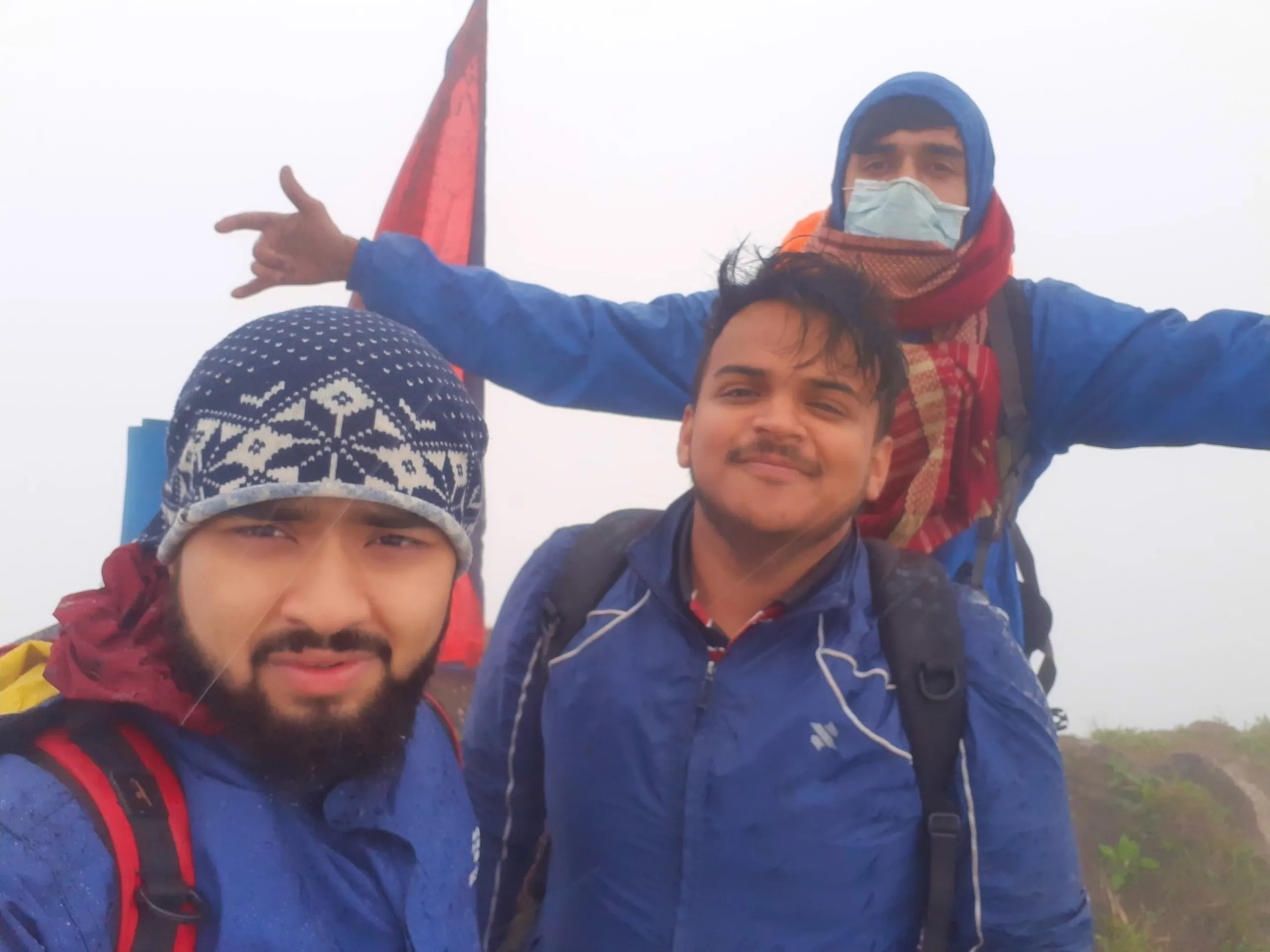 Group photo of three young men posing together at the foggy Bethanchowk summit near the red Nepali national flag visible in the upper left corner. The man in front wears a navy and white patterned beanie and blue jacket with red and maroon scarf, while the man beside him in a blue jacket with white piping smiles with a light mustache. Behind them, a third friend wearing a blue hooded jacket, navy cap, and red checkered scarf covering his face with a surgical mask underneath makes a hand gesture with arm extended. Thick white fog fills the entire background. This captures a memorable trio photo at Bethanchowk flag summit, Nepal Himalayan trekking group photography, misty mountain peak celebration, friends adventure memories Kathmandu hills.