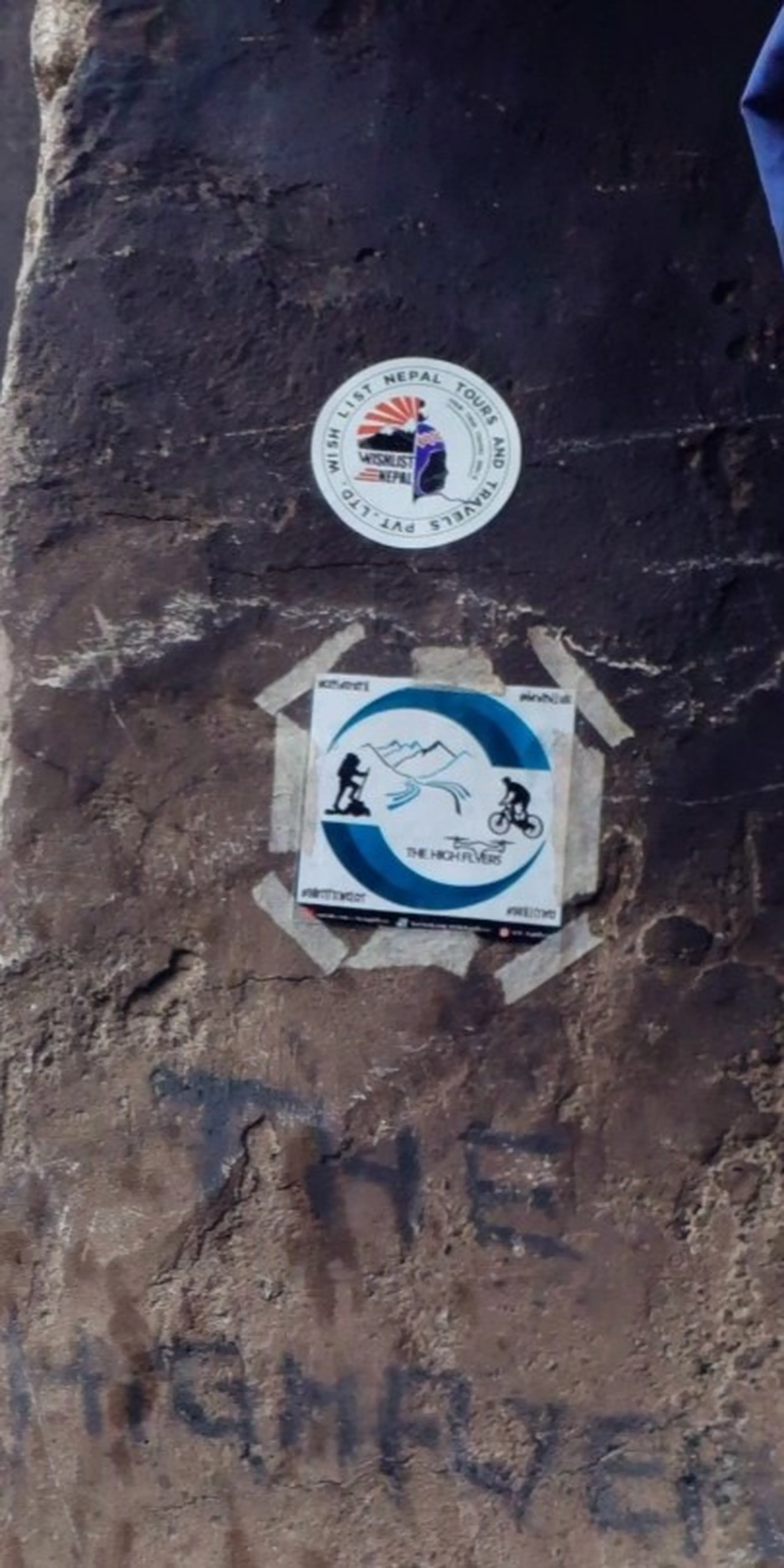 Close-up photo of two circular promotional stickers affixed to a dark weathered rock surface at Bethanchowk summit, with handwritten graffiti reading "THE HIGH FLYER" visible below in black marker. The top sticker shows a "Wish List Nepal Tours and Travels" logo with a stylized mountain and sun design in orange, blue, and white colors, while the bottom square sticker displays "The High Flyer" branding featuring icons of a hiker, mountains, and a cyclist in blue and white circular design. This documents trekking company branding and signage left at Bethanchowk highest point, Nepal adventure tourism marketing, Himalayan trekking company stickers, mountain summit landmarks Kathmandu hills.