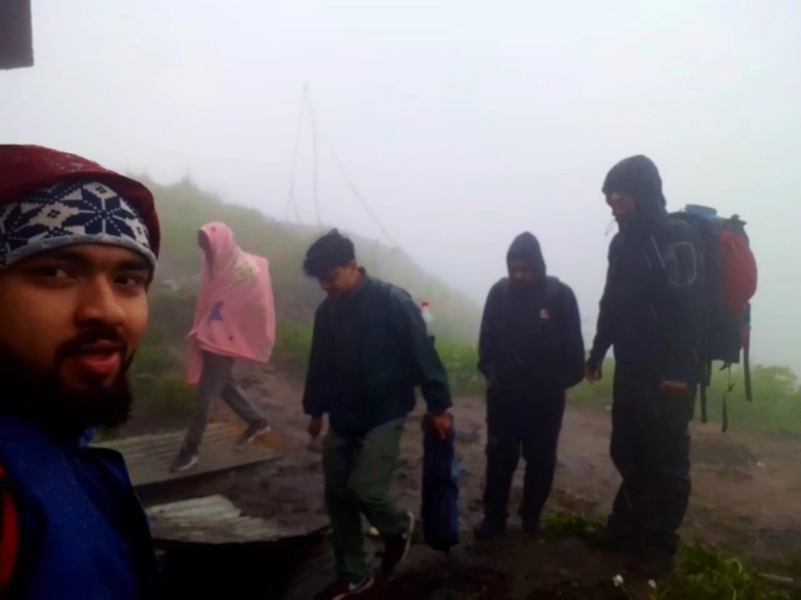 Atmospheric group photo taken in heavy rain and fog showing a bearded young man in a maroon and navy patterned beanie and blue jacket with red backpack straps visible in the foreground, while behind him four other hikers walk along a muddy path near a corrugated metal structure. One person wears a bright pink rain poncho with hood up, another carries a blue rolled item, and two others wear dark hooded rain jackets with one carrying a large red and grey backpack with rolled sleeping mat. Thick fog and visible rainfall create a challenging weather atmosphere. This captures the difficult rainy descent from Bethanchowk highest point, Nepal monsoon trekking challenges, Himalayan rain hiking adventure, group trekking in bad weather Kathmandu hills.