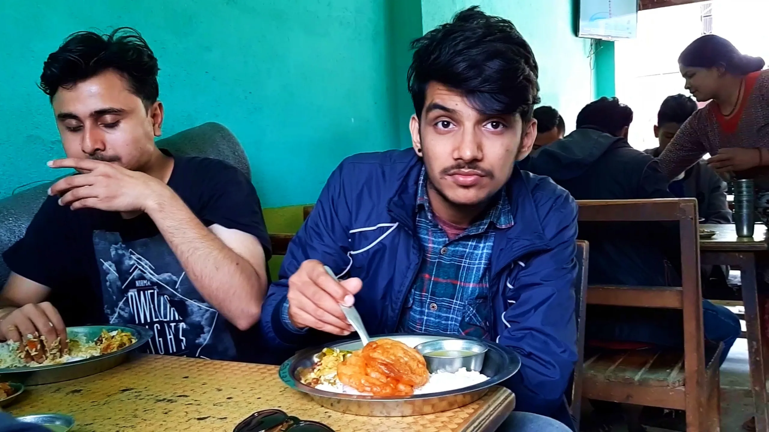 Indoor restaurant scene showing two young men seated at a wooden table eating a traditional Nepali dal bhat meal during a road trip stop. The man in the foreground wears a navy blue jacket over a blue plaid shirt, holding a spoon over a steel plate filled with white rice, spicy orange-red marinated chicken curry pieces, sautéed green vegetables, and a small bowl of clear soup or achar, looking directly at the camera with a neutral expression and sunglasses placed on the table. Beside him, another man in a black "Dwelling Nights" graphic t-shirt eats with his hand, looking down at his plate of rice and curry. The restaurant has turquoise green painted walls, a wall-mounted TV screen, wooden chairs and benches, with other diners visible in the background including a woman in a red top. This represents authentic Nepali dal bhat lunch stop, local Nepal restaurant food, road trip dining Kathmandu to Bethanchowk, Nepali cuisine travel experience.