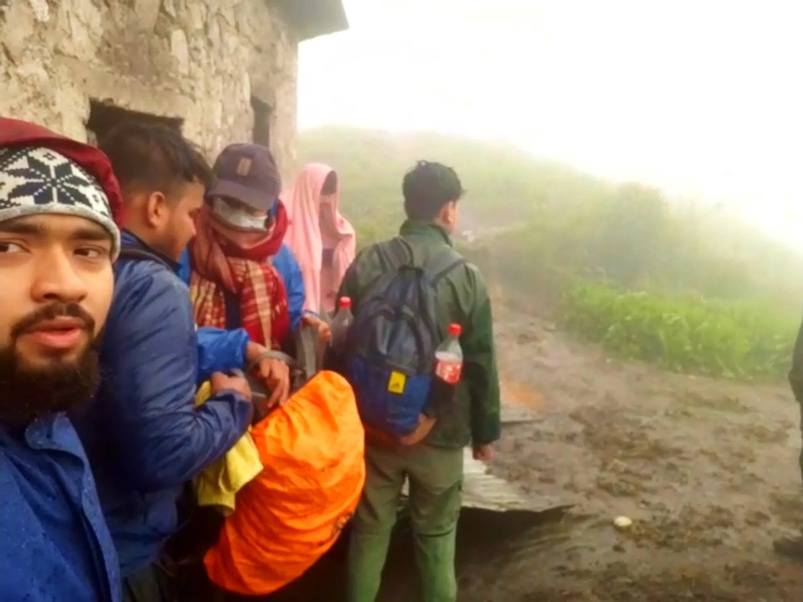 Photo taken in rainy foggy conditions showing a group of hikers standing near the corner of an old stone building at Bethanchowk, with a bearded young man in a maroon and navy beanie and blue jacket in the foreground close to the camera. Behind him, friends wear navy cap, pink rain poncho, and dark hooded jackets, with one person holding an orange stuffed sack or duffel bag and another carrying a navy backpack with a red soda bottle attached and a blue accessory clipped to the side. Wet muddy ground and bright overcast fog dominate the scene with green vegetation visible on the right. This captures the group preparing to depart Bethanchowk highest point in heavy rain, Nepal monsoon trekking departure, Himalayan rainy weather hiking, adventure group travel Kathmandu hills.