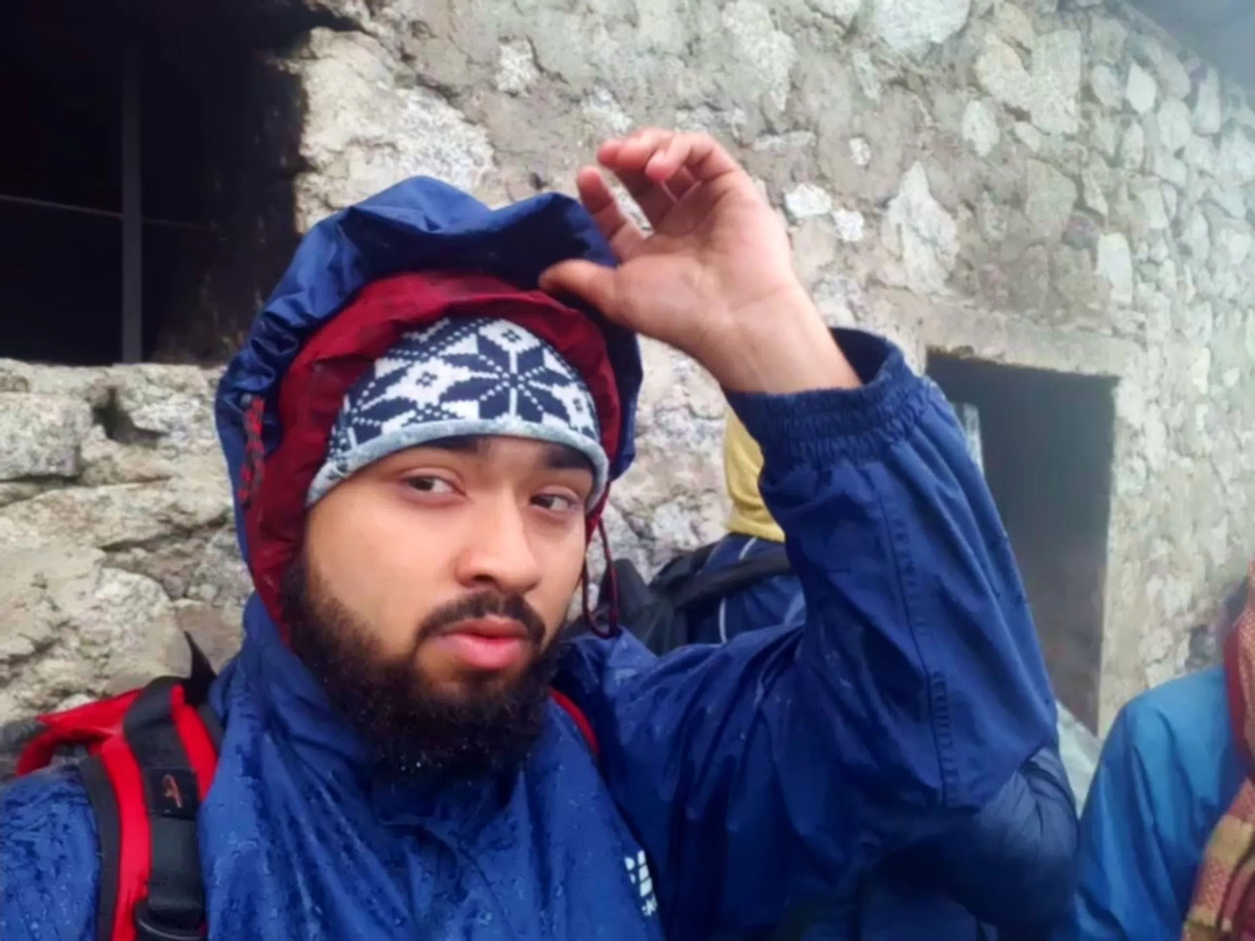 Close-up selfie of a bearded young man wearing a maroon and navy patterned knit beanie under a navy blue rain jacket hood, raising one hand to adjust his hood while standing in front of the weathered grey stone wall of the old cottage at Bethanchowk, with a red backpack visible on his shoulder. In the background, another hiker wearing a yellow garment and another in a blue jacket with red checkered scarf are partially visible against the stone building's dark window openings. This captures a rainy-day portrait at the historic stone shelter Bethanchowk highest point, Nepal Himalayan monsoon trekking gear, rainy mountain hiking adventure, group trekking memories Kathmandu hills.