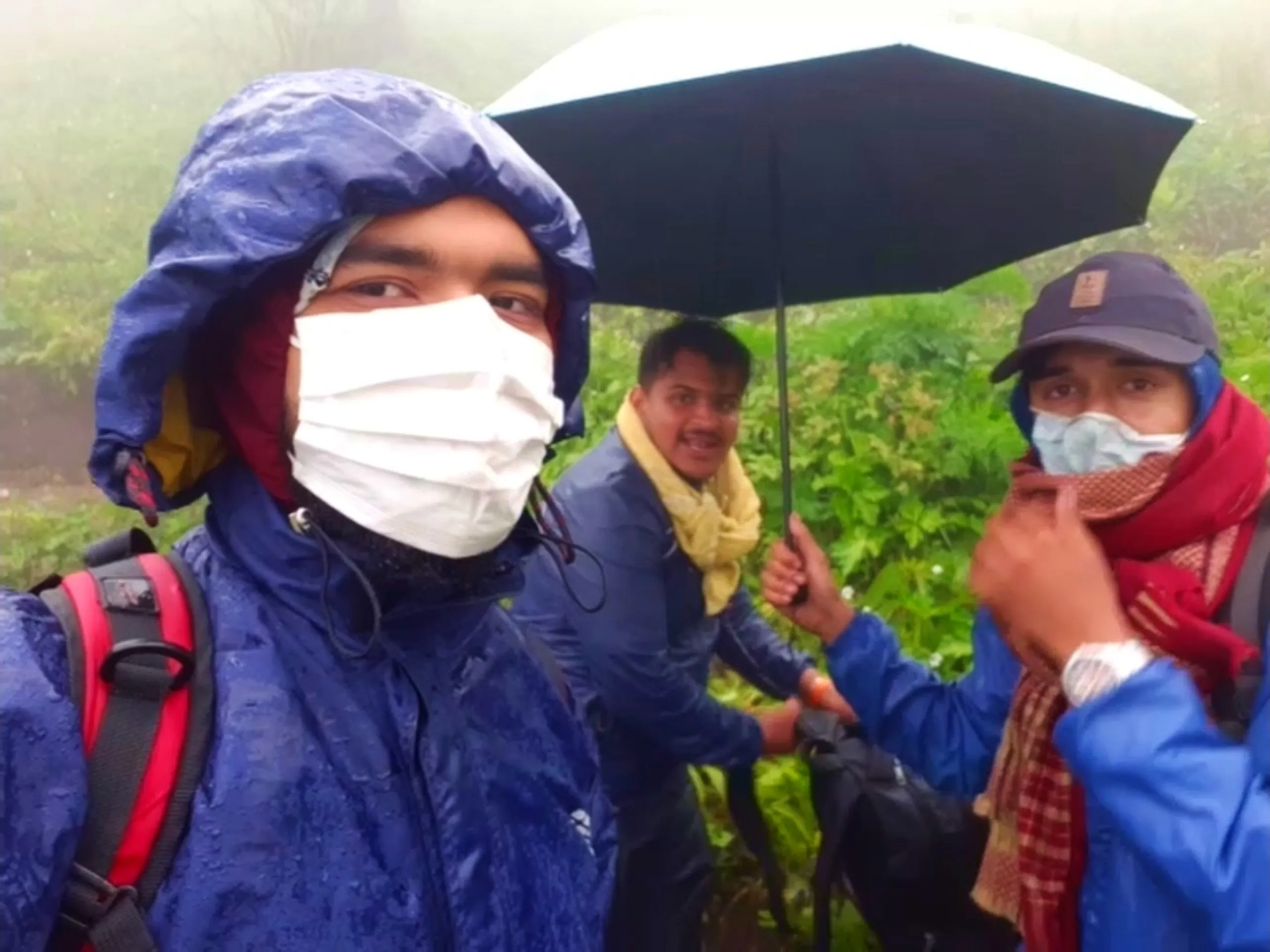 Group photo of three young hikers in heavy rain near a green bushy area, all wearing surgical face masks for protection. The man on the left wears a navy blue rain jacket with hood up and red backpack straps, while the man in the center wears a blue rain jacket with yellow scarf, smiling and holding a black umbrella overhead. On the right, another hiker wearing a navy cap and blue rain jacket with red checkered scarf adjusts his face mask, looking at the camera. Wet green foliage fills the background. This captures friends braving heavy monsoon rain during Bethanchowk trekking trip, Nepal rainy season hiking, COVID-era outdoor adventure, group trekking in bad weather Kathmandu hills.