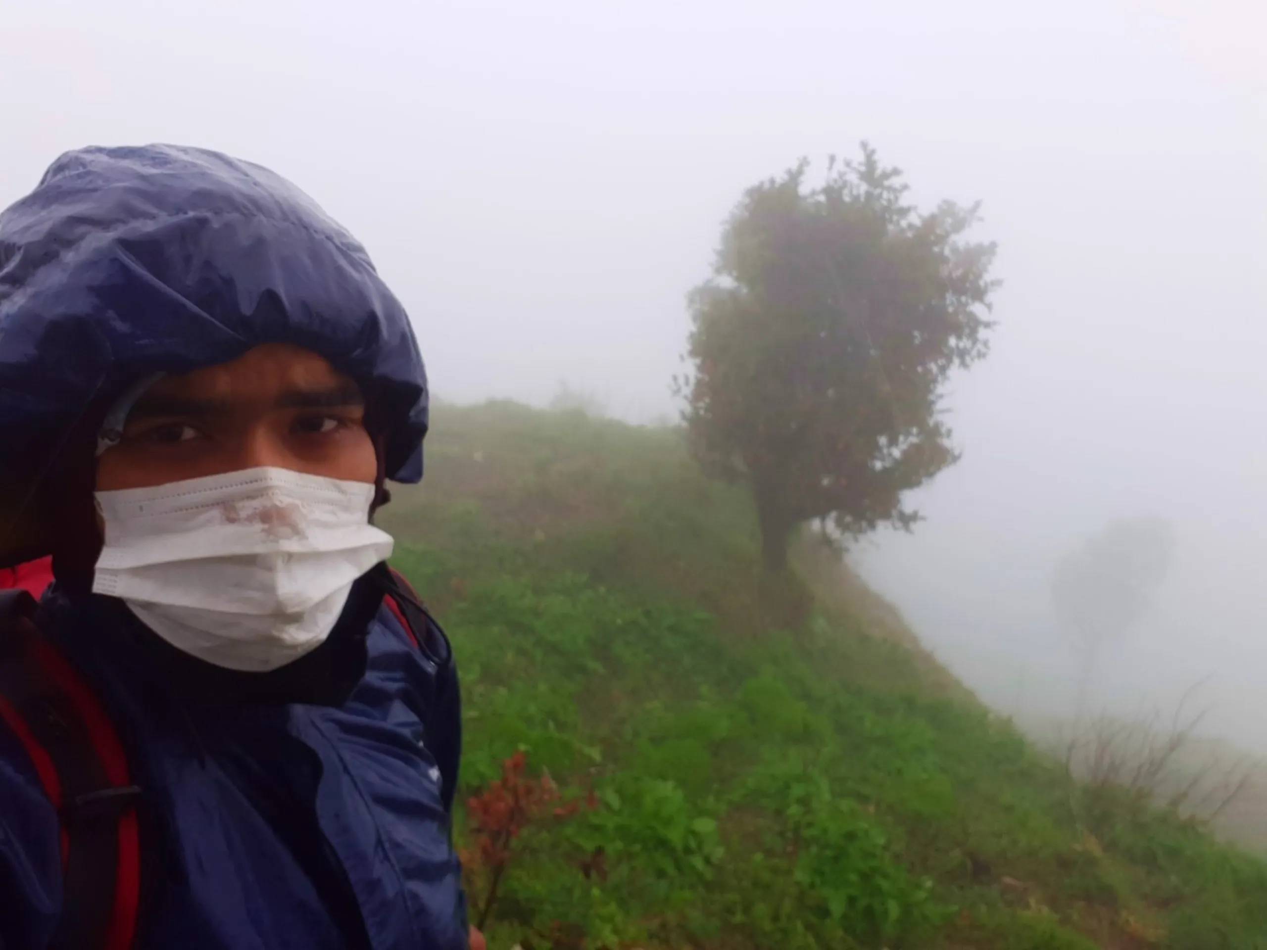 Close-up selfie of a young man wearing a navy blue rain jacket hood and a white surgical face mask, looking seriously at the camera with a red backpack strap visible, standing on a misty green hillside. In the background, a lone tree with dark green foliage stands silhouetted against thick white fog on a grassy ridge, with another faint tree shape visible further in the distance through the mist. This captures a masked rainy hike portrait at Bethanchowk highest point, Nepal monsoon trekking safety gear, foggy mountain ridge hiking, adventure travel during pandemic Kathmandu hills.