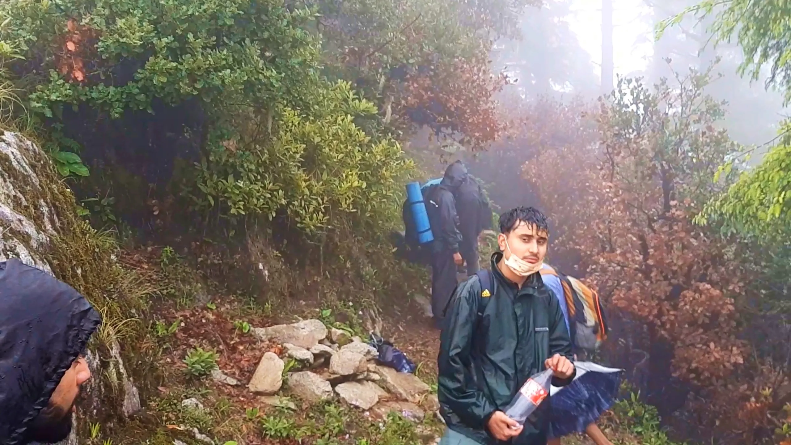 Photo of three hikers walking along a misty forest trail covered in reddish-brown autumn leaves and surrounded by moss-covered rocks and green-and-orange foliage. In the foreground, a young man wearing a dark green Adidas-branded rain jacket and white surgical face mask holds a plastic water bottle and a blue rain item, looking at the camera. Behind him, another hiker in dark rain gear carries a blue rolled sleeping mat on his backpack, while a hand wearing a dark rain sleeve is visible at the left edge of the frame. Bright misty light filters through the autumn-colored forest canopy. This captures the colorful autumn forest trail during Bethanchowk descent, Nepal Himalayan rainy forest hiking, monsoon trekking group photo, scenic foliage trail Kathmandu hills.