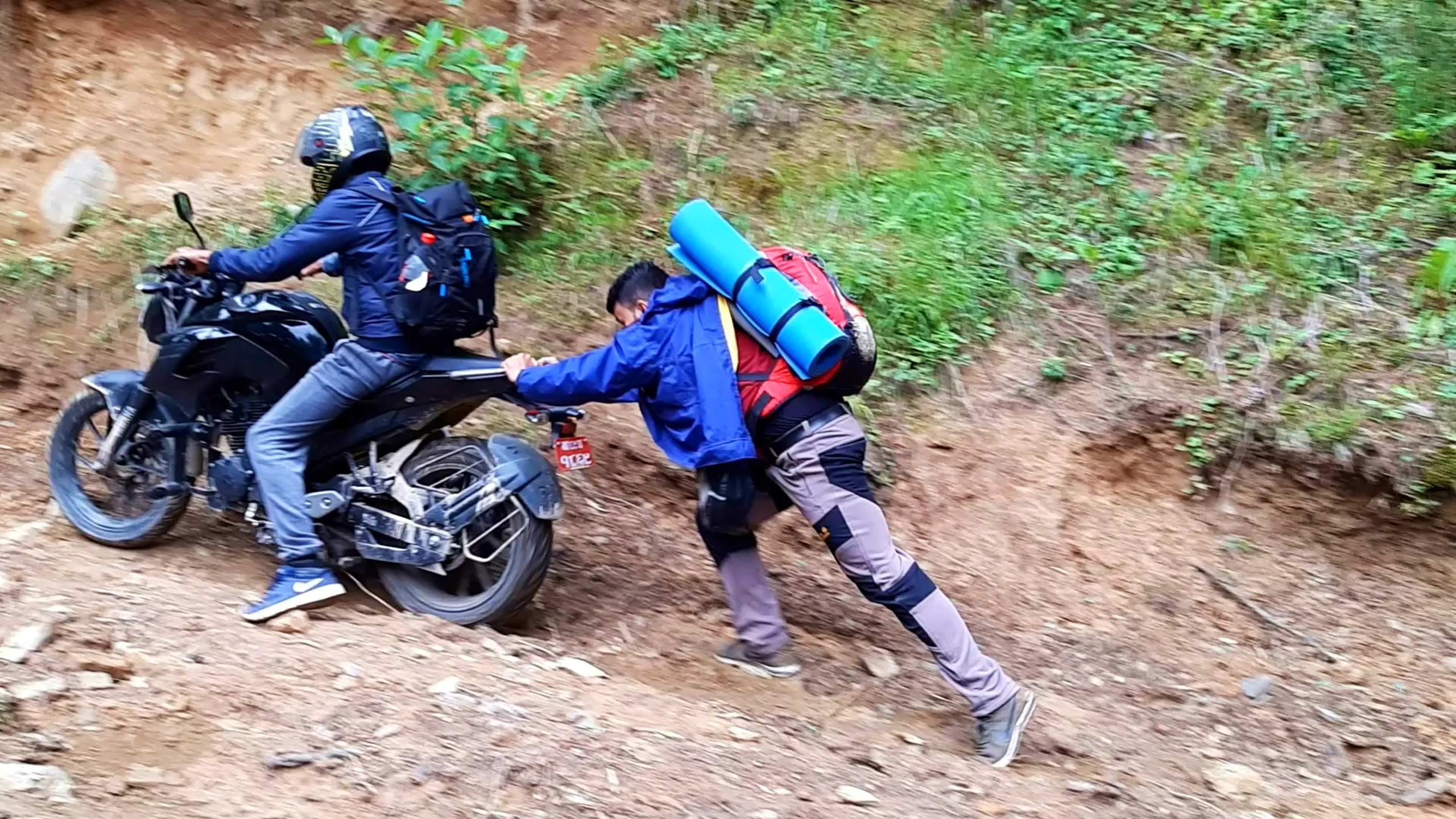 Action photo of a motorbike struggling on a steep muddy off-road dirt trail surrounded by dense green forest vegetation, with one rider in a black helmet and navy jacket with a black backpack seated on the bike attempting to ride up the slope, while a second hiker wearing a blue rain jacket, grey cargo pants, a red backpack with a turquoise rolled sleeping mat strapped on top, and a black helmet pushes the rear of the motorbike for momentum. The motorbike's rear wheel kicks up loose dirt and stones on the steep reddish-brown earthen incline. Red Nepali number plate visible on the bike. This scene depicts the challenging off-road motorbike trekking route to Bethanchowk, extreme adventure riding Nepal hills, monsoon trail riding Himalayas, off-road biking adventure Kathmandu.