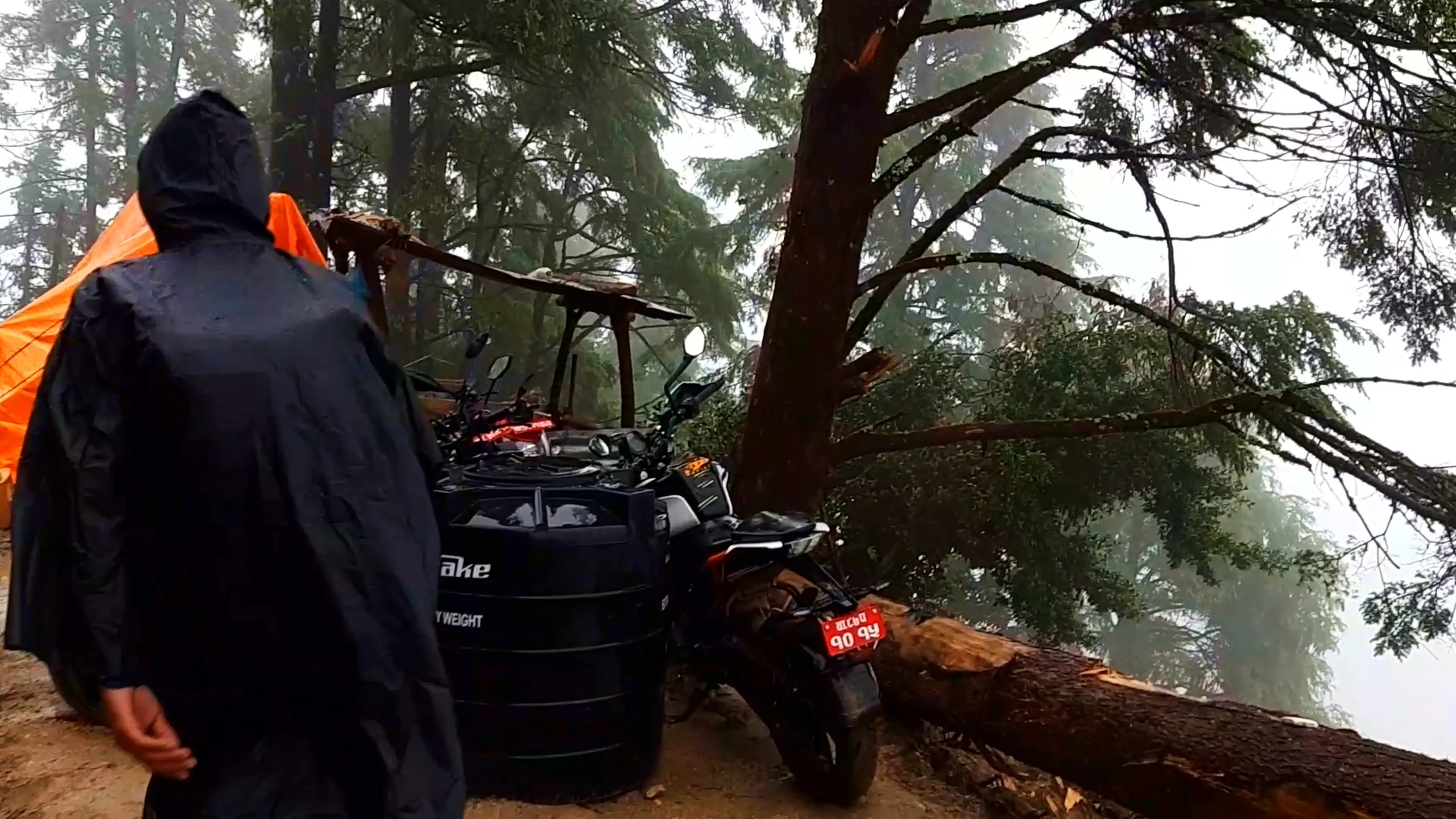 Atmospheric photo of a hiker wearing a dark hooded rain jacket walking past a parked white and black KTM Duke motorcycle and a large black water storage tank near an orange camping tent, under tall pine trees with a fallen log lying across the misty forest clearing. The motorcycle has a red Nepali number plate and rearview mirrors visible, with thick fog filling the background between the trees. This shows the motorbike camping spot along the Bethanchowk trekking route, Nepal motorbike adventure camping, Himalayan forest campsite, off-road trekking gear Kathmandu hills.