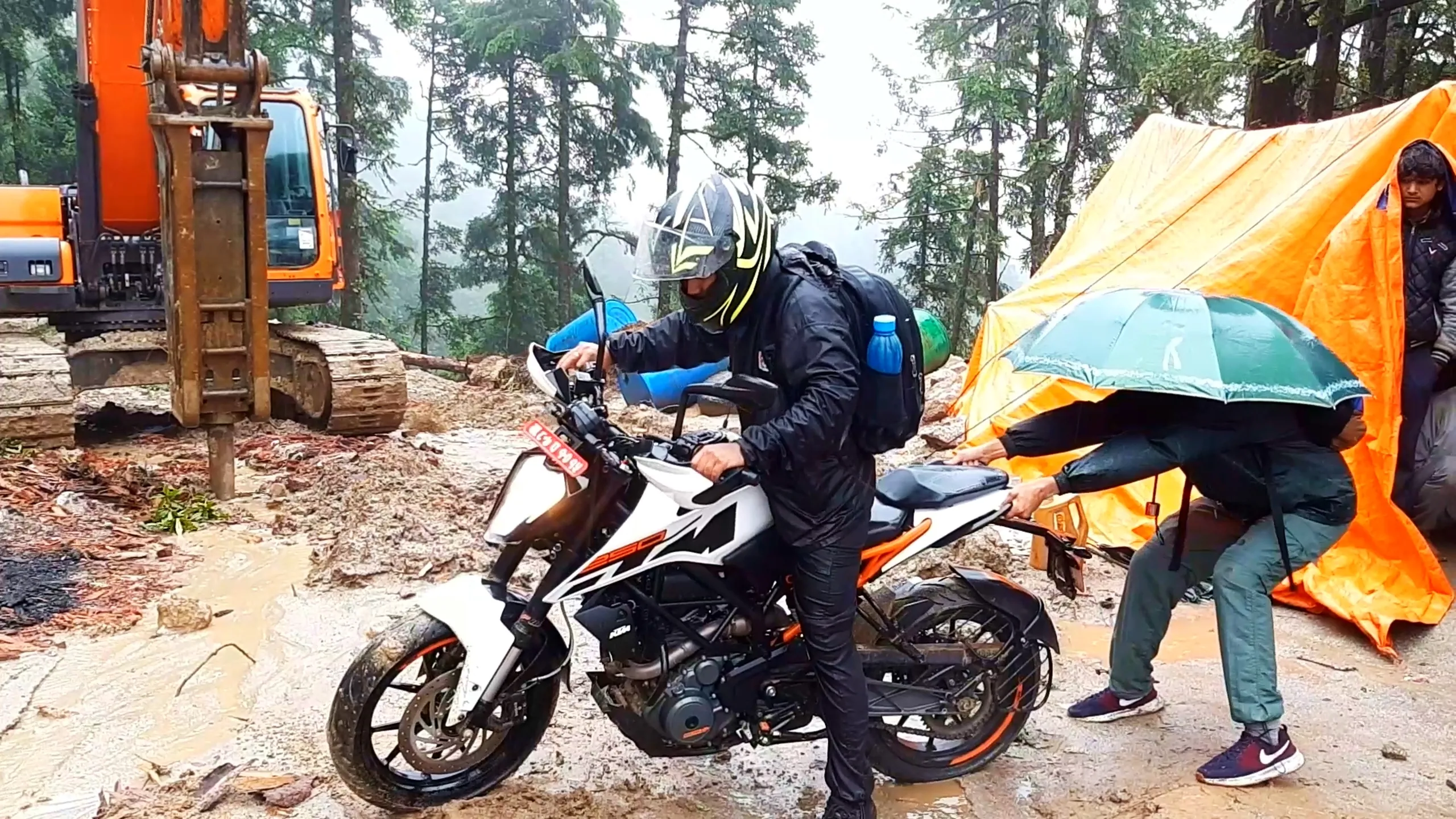 Action photo of a man wearing a black and yellow graphic motorcycle helmet, black riding jacket, and grey backpack with water bottle, mounting a white and black KTM Duke 250 motorcycle with orange accents and a red Nepali number plate, parked on a muddy clearing near excavator construction machinery. Beside him, another hiker holding a green umbrella reaches toward the motorbike's rear seat, with an orange tent visible on the right and another person partially visible near the tent entrance. Tall pine trees and grey overcast sky fill the background. This captures preparing to ride out from the Bethanchowk forest campsite, Nepal motorbike trekking departure, Himalayan adventure motorcycle gear, off-road KTM Duke trekking Kathmandu hills.