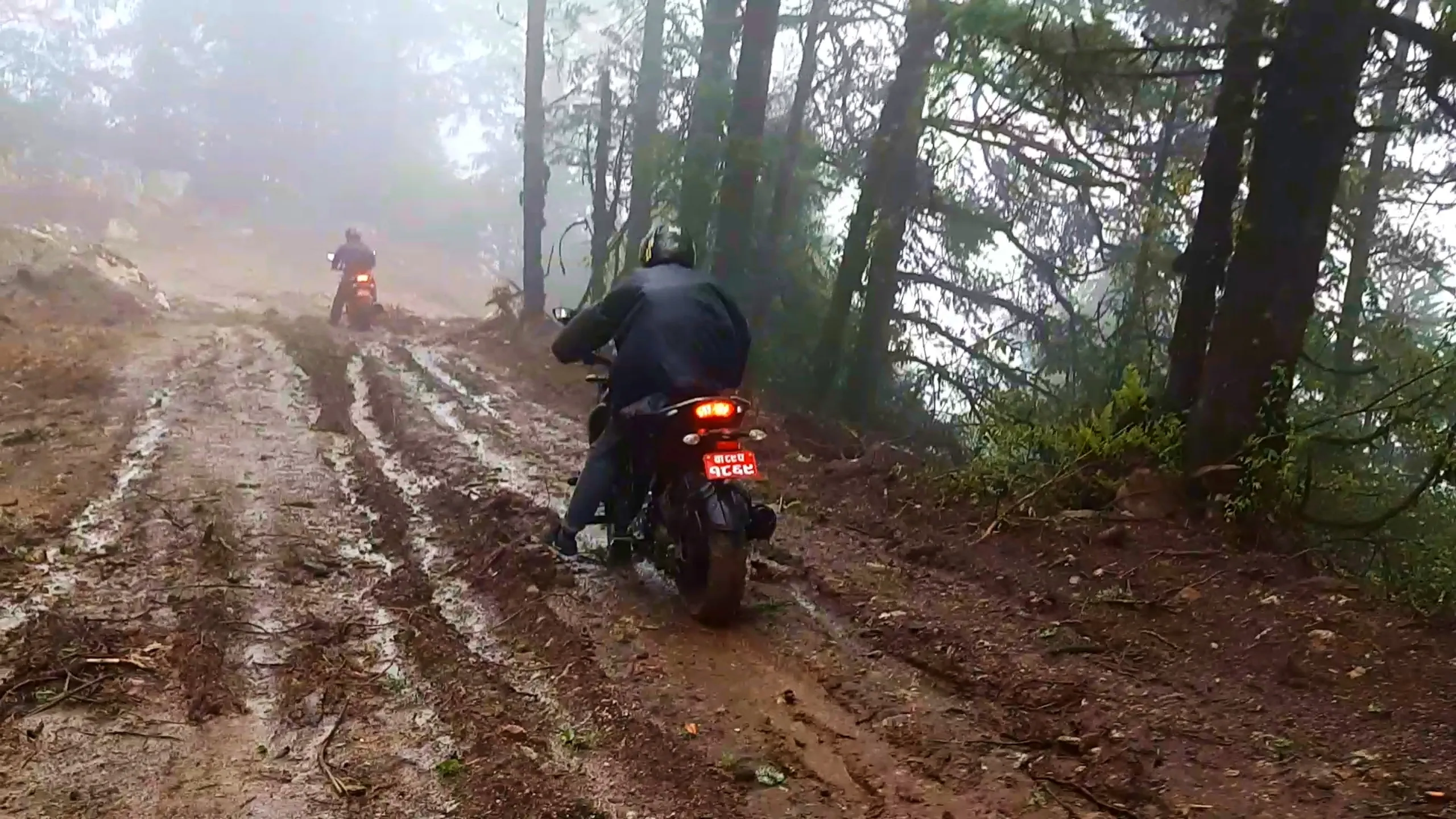 Action photo taken from behind showing two motorcyclists riding away on muddy forest trails covered in wet mud and water puddles, with the nearer rider wearing a dark helmet and jacket on a black motorbike with red taillight illuminated and red Nepali number plate, while another rider on a similar bike is visible further ahead navigating the muddy uphill path. Tall pine and cedar trees with hazy fog line both sides of the trail. This captures the muddy motorbike trail ride away from Bethanchowk, Nepal off-road monsoon motorcycling, Himalayan forest trail riding, adventure motorbike trekking Kathmandu hills.