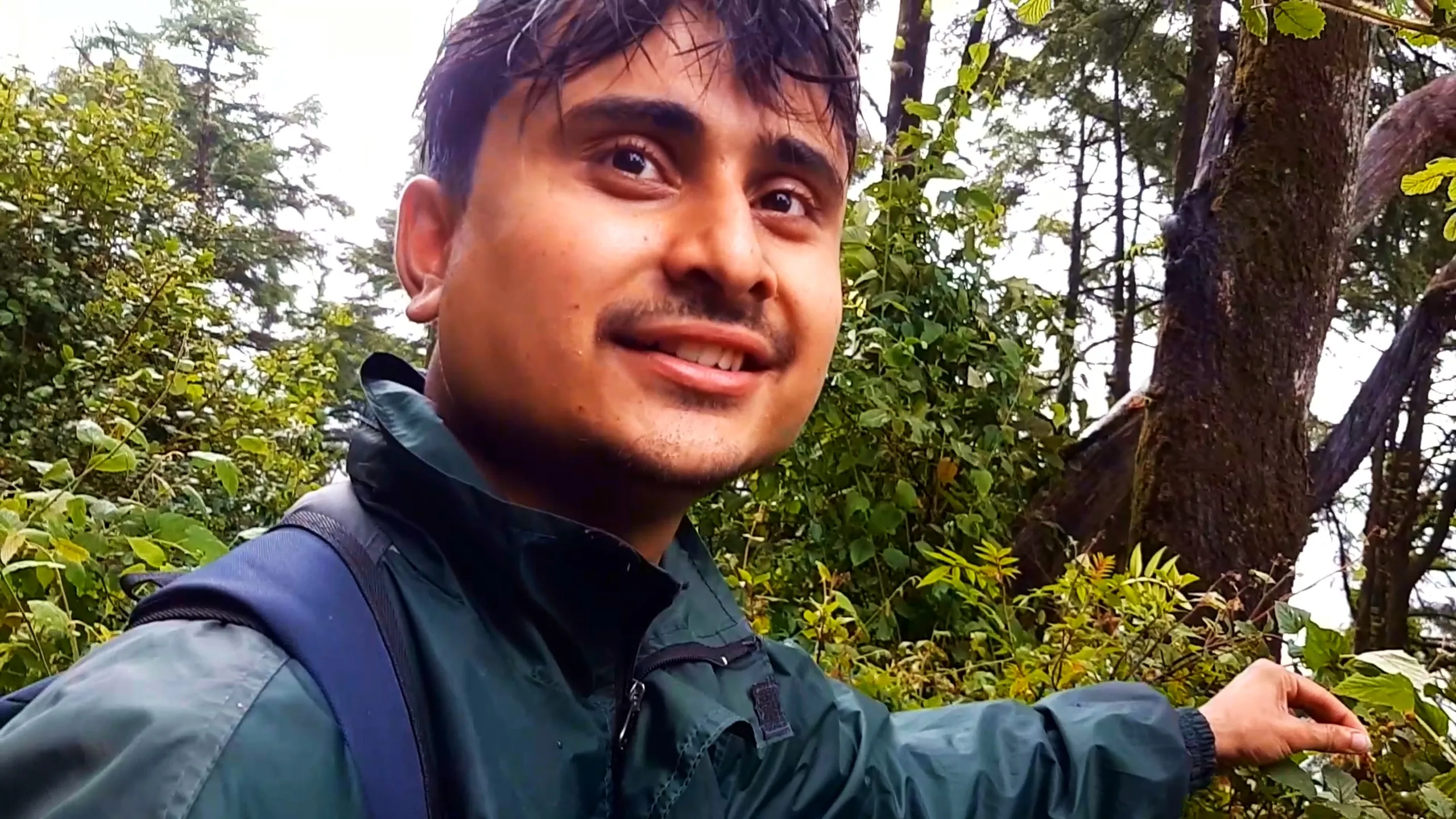 Close-up selfie of a smiling young man wearing a dark green rain jacket with a navy blue backpack strap, reaching out with one hand toward green forest foliage, with tall pine trees and bright misty sky in the background. This captures a cheerful moment foraging in the forest during Bethanchowk trekking descent, Nepal Himalayan forest exploration, nature trekking adventure, group hiking memories Kathmandu hills.