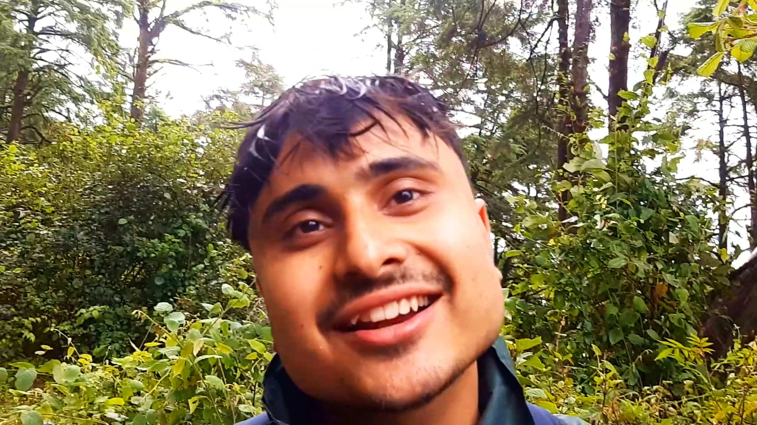 Close-up selfie of a smiling young man with damp tousled dark hair, wearing a dark green rain jacket, looking at the camera with a warm smile against a backdrop of dense green forest foliage and tall pine trees with bright overcast sky. This captures a happy candid moment during the Bethanchowk forest trekking descent, Nepal Himalayan adventure travel photography, rainy forest hiking portrait, group trekking memories Kathmandu hills.