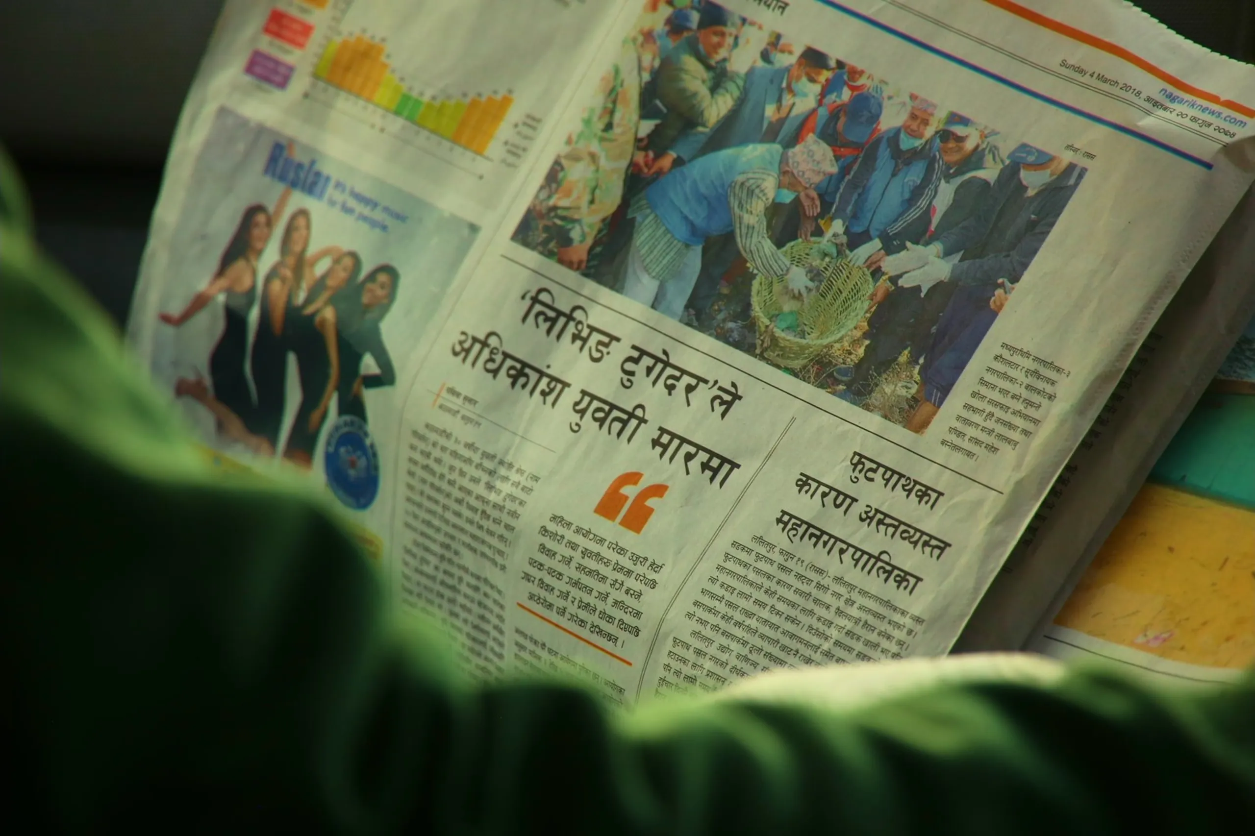 A candid, slightly blurred close-up of someone reading a Nepali language newspaper, held open at what appears to be the front or a main news page. The reader - visible only as a pair of hands and a green-clothed torso - is absorbed in the content. The newspaper is printed in Devanagari script, with a large headline text in bold black and a photograph on the page showing a group of people at some kind of official or public event - figures in formal or traditional dress gathered together. A small advertisement with an image of women is visible in the upper left corner of the page. The image is shot at close range with a shallow depth of field, so the text is partially readable but the overall impression is of a quiet, ordinary moment of reading the news - perhaps on a bus or in a waiting room.