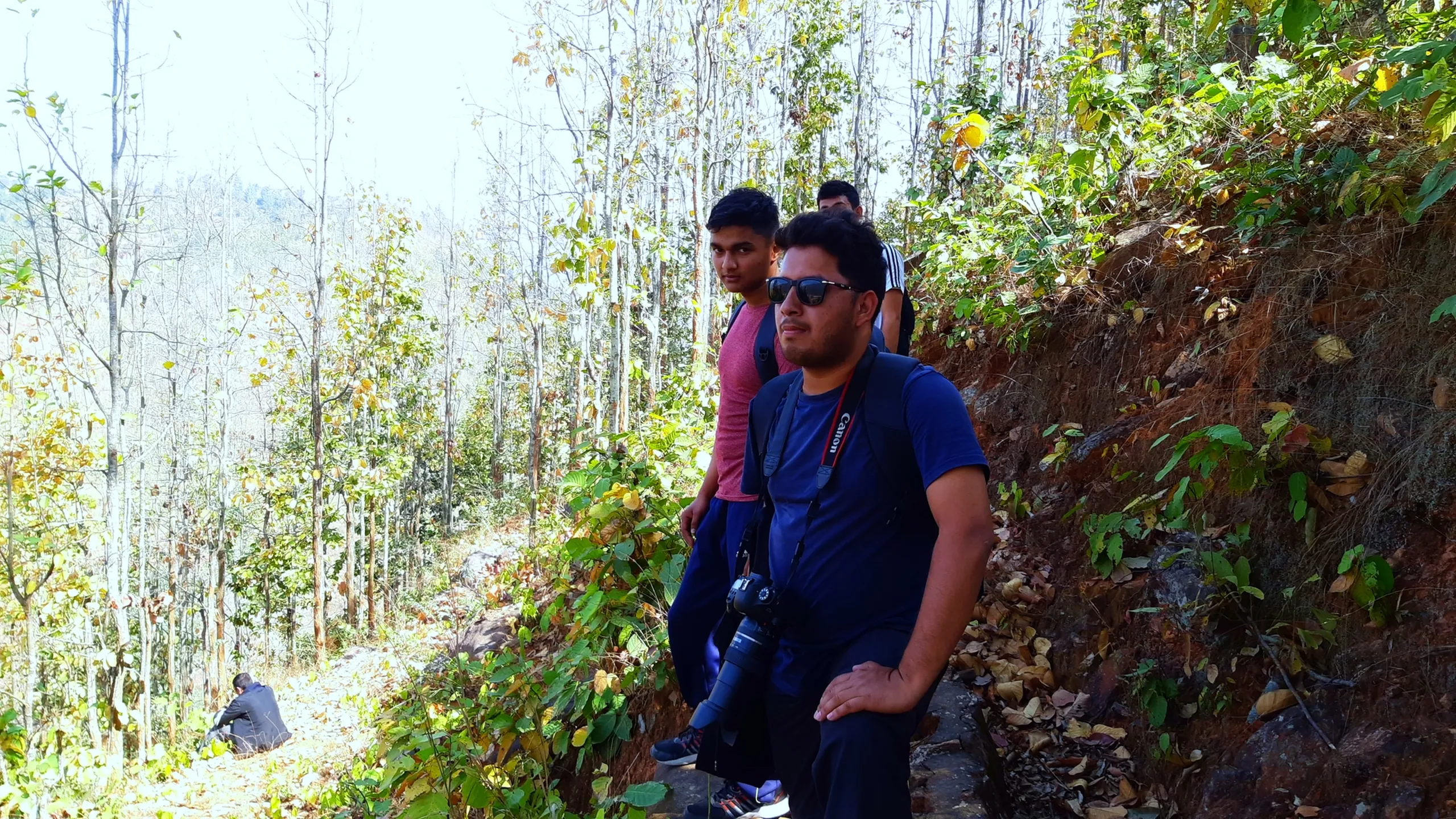 Two men pause on a hillside trail among deciduous trees with yellowing autumn leaves, both looking outward at the view rather than directly at the camera. The man in the foreground is more prominent - he wears dark sunglasses and a dark navy or charcoal top, with a camera hanging around his neck, suggesting he is the trip photographer. His expression is relaxed and contemplative. Behind him, slightly further up the slope, the second man wears a pink or light red top. The trees around them are tall and slender, their leaves turning yellow-green in the autumn light that filters through. The ground is dry and sloped. The image captures a quiet pause on the trail - two people standing still in the moving world of the forest.