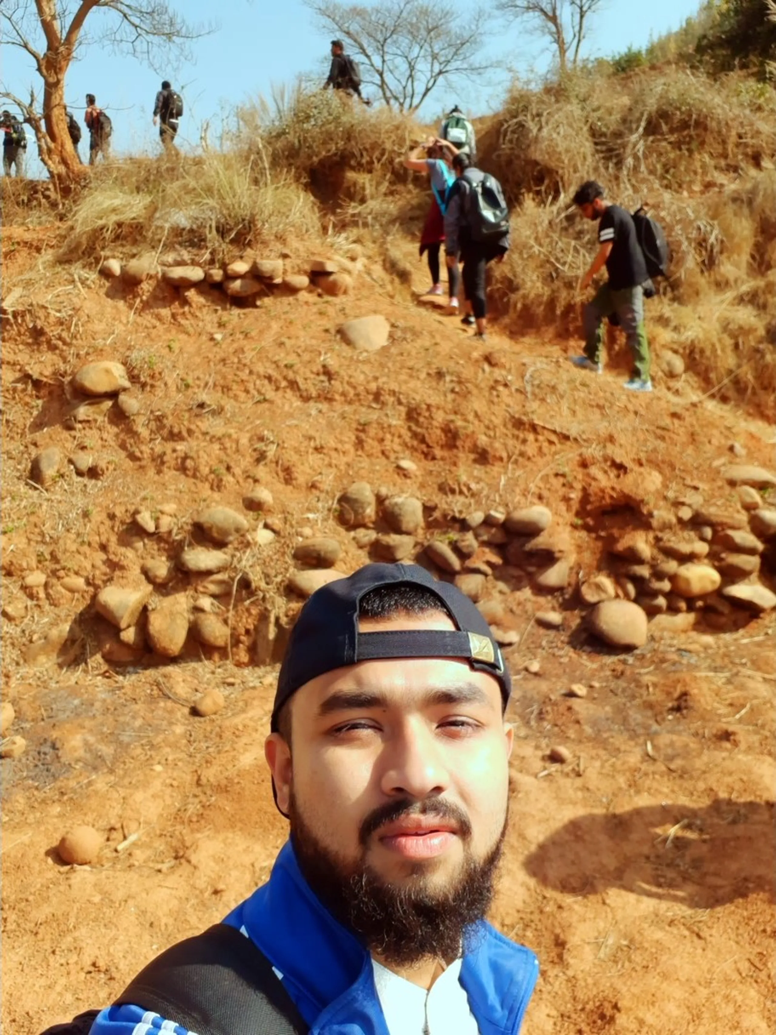 A trail selfie taken on a steep, rocky hillside of vivid red-orange earth. The same bearded man with the backwards navy cap and blue jacket from the opening image takes the selfie in the close foreground, one eye slightly squinted - mid-action, casual. Behind him, the trail rises steeply and dramatically. Several other hikers are visible climbing the slope - three or four figures spread across the red-earth incline, their backs to the camera. The ground is rough red laterite earth dotted with rounded grey and white smooth boulders scattered across the slope like oversized marbles. The path is rugged and uneven. Dry scrub vegetation borders the trail at the top of the frame. The image conveys effort, camaraderie, and a trail that demands real climbing.