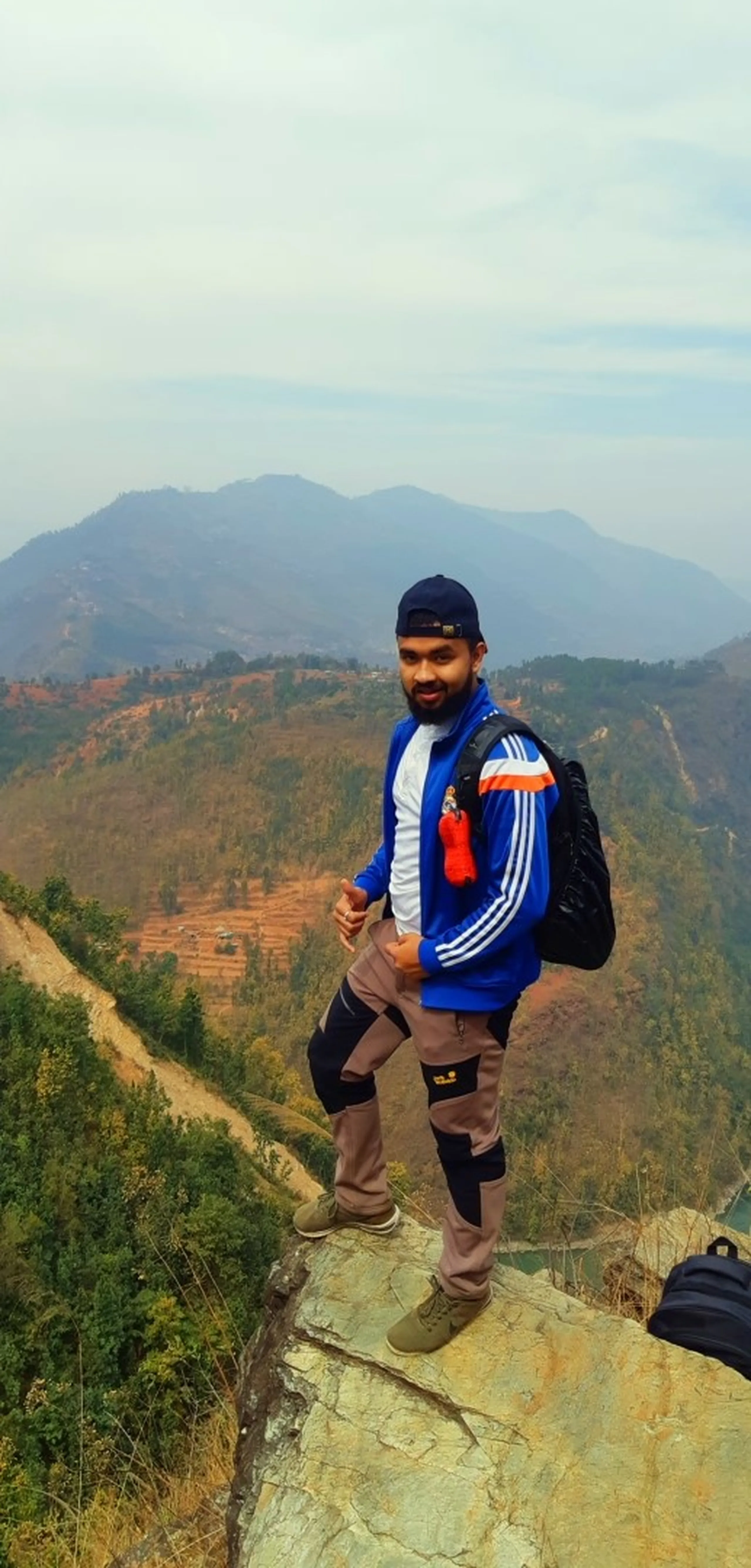 A triumphant summit portrait. A young man stands on a large, flat rock at a high-elevation viewpoint, posing with relaxed confidence and a wide, genuine smile. He wears a blue Adidas-style jacket with white stripes down the sleeves, grey-khaki trekking trousers with a visible knee brace or support strap on one knee (suggesting a real trekker who takes the effort seriously), a backwards dark cap, and carries a black backpack. He faces the camera squarely, one foot slightly forward, body open. Behind him stretches a sweeping panoramic view of layered mountain ridges receding into the haze - slopes patchy with terraced fields and scrub, valley floors barely visible far below, and a pale hazy sky above. The image is the definition of hard-earned joy at altitude.