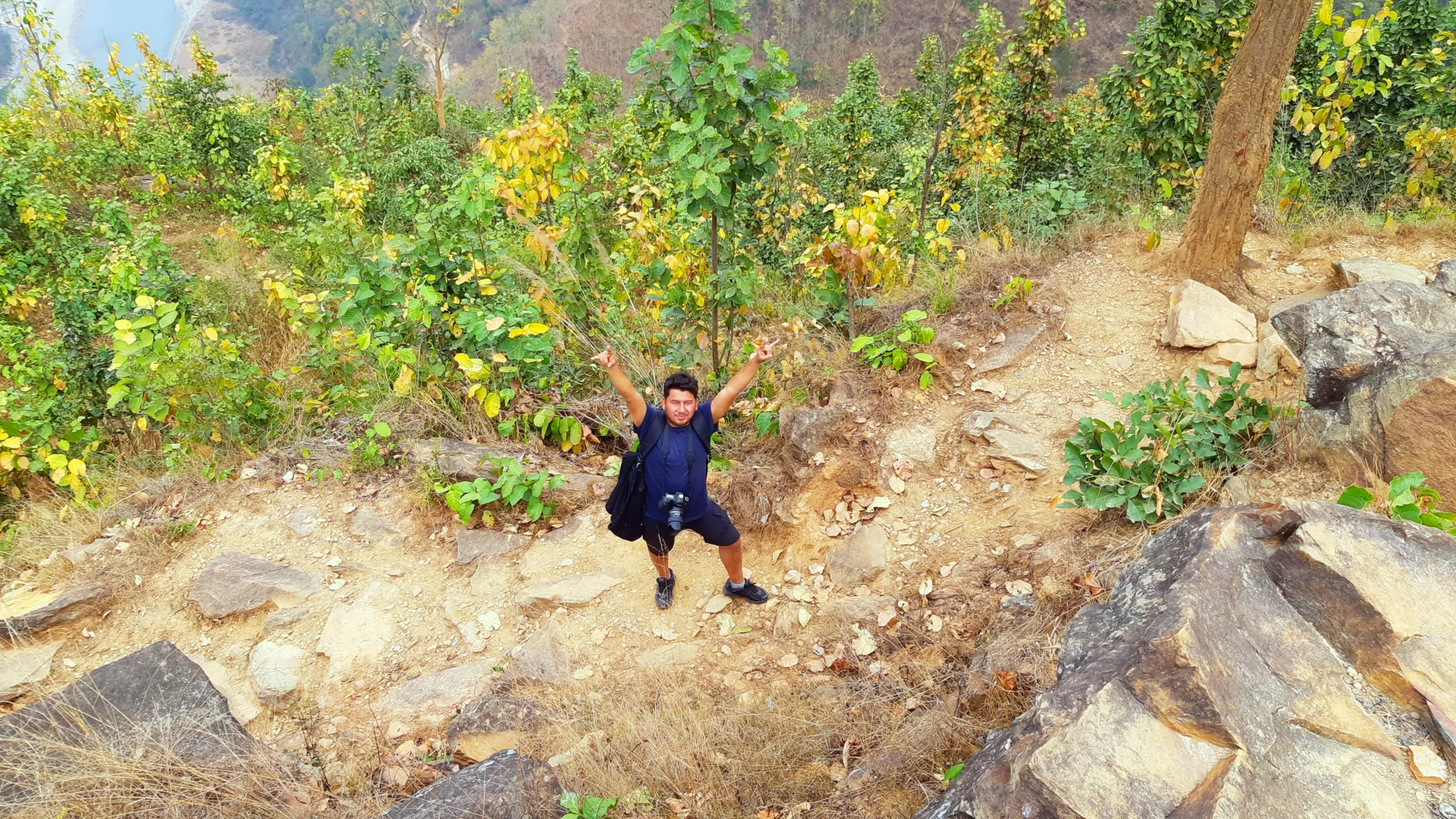 A joyful, top-down aerial or elevated photograph of a man on a rocky hillside, shot from directly above or from a steep angle above him. He stands among rocks and small trees and shrubs with vivid golden-yellow autumn leaves, and he has spread both arms wide apart - a gesture of pure freedom, as if about to take flight or simply surrendering to the joy of being in the mountains. His body is small in the frame relative to the rocks and foliage surrounding him. The warm yellow and orange of the autumn leaves, the grey-brown of the rocks, and the figure with outstretched arms together create a playful, exuberant image full of energy and lightness.