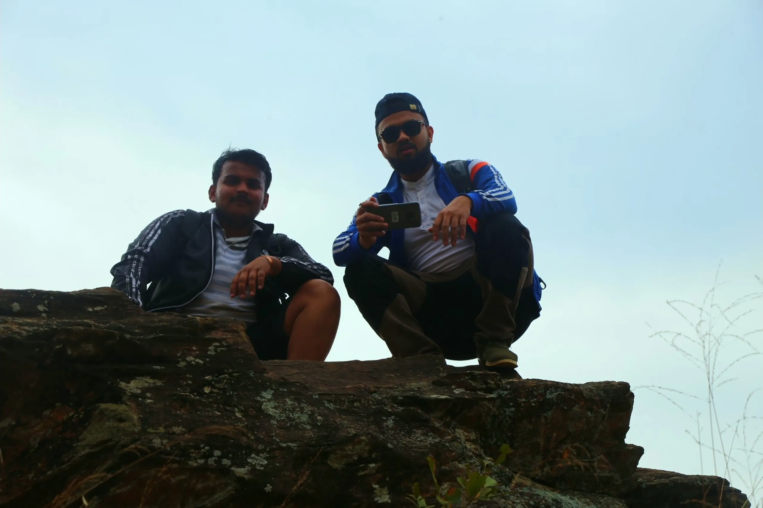 Two men sit together on top of a large, flat grey rock at what appears to be a high summit or exposed ridge, silhouetted slightly against a pale, overcast grey-white sky. The light is flat and slightly backlit, making their faces shadowed and the overall image slightly underexposed. One man sits more loosely, arms relaxed. The other squats with his knees bent, holding what appears to be a phone or camera. Both wear casual outdoor jackets. A thin, bare tree branch extends into the upper right of the frame. The mood is quiet and end-of-day - two friends resting at the top of something they climbed, the world grey and wide behind them.