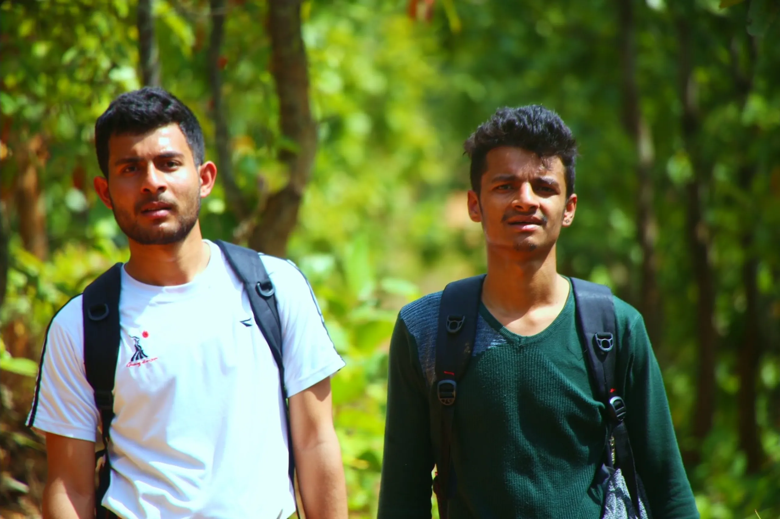 A clean, close-up portrait of two young men standing side by side in a lush green forest, both facing the camera directly. Both carry backpacks visible on their shoulders. The man on the left wears a white t-shirt with a small graphic or logo, and has a lean, calm face with dark eyes and short hair. The man on the right wears a dark forest green t-shirt and has a slightly broader face with a similarly composed, direct expression. Neither smiles broadly - their looks are quiet, natural, and candid. The background behind them is completely filled with soft, blurred green forest bokeh - lush, bright, and deep. The image has the quality of a natural, unforced friendship portrait.