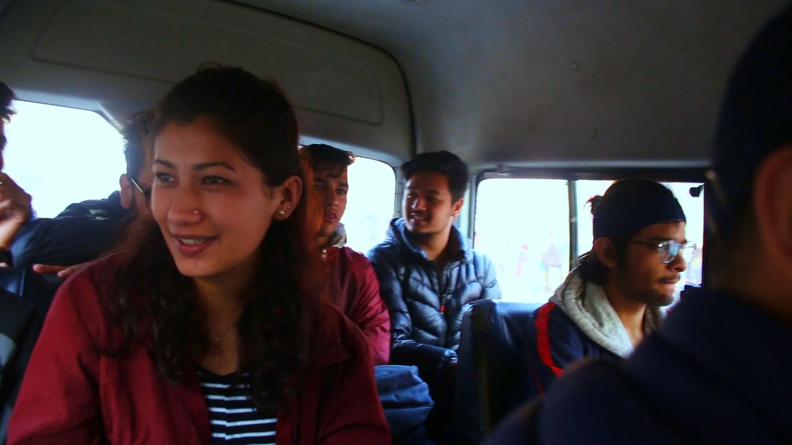 The interior of a crowded minibus or shared van, packed with passengers making a journey through what is likely a Nepali town or city. The seats are old and close together. In the center of the frame, a young woman is the focal point - she wears a bold red jacket over a navy and white striped shirt, and she is smiling openly and warmly, her expression bright and relaxed. Around her, several other passengers - mostly young men - are dressed in winter jackets and dark clothing, sitting in various directions. The van's interior is plain and functional - no decoration, simple fabric seats, natural light coming from windows behind. The scene feels like the beginning of an adventure - a group of people in transit together, the woman's smile the warmest thing in the frame.