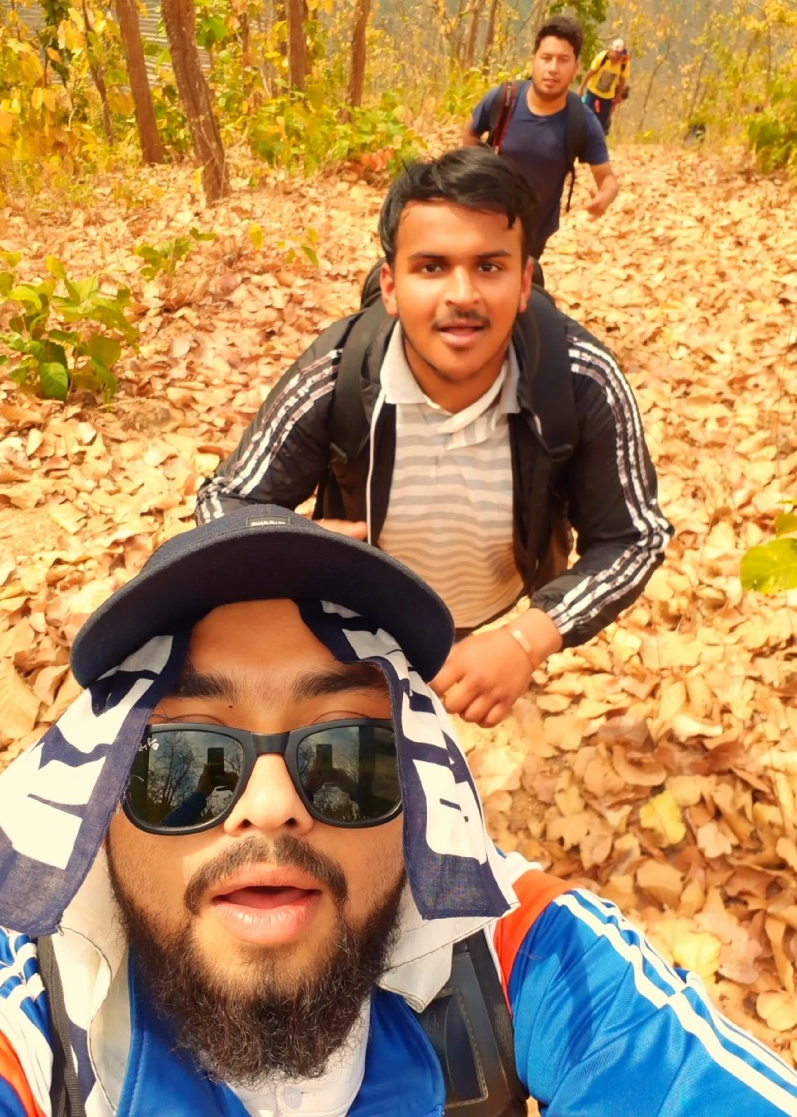A lively trekking selfie taken on a forest trail carpeted entirely in dry, fallen autumn leaves of deep orange and brown. In the sharp foreground, a young bearded man with a neatly trimmed beard takes the selfie - he wears dark reflective sunglasses, a backwards navy blue cap, and a blue jacket with white accents, and carries a backpack. His mouth is slightly open, mid-sentence or mid-laugh. Directly behind him, a second young man in a grey-striped collared shirt and a dark track jacket also carries a backpack, looking toward the camera with a calm, friendly expression. Further back up the trail, a third figure - slightly out of focus - walks with a bag on their back. The forest around them is dry and deciduous, the trees bare or thinly leaved, the ground entirely obscured by a thick blanket of crisp orange-brown leaves. The group is clearly mid-trek on a bright, cool day.