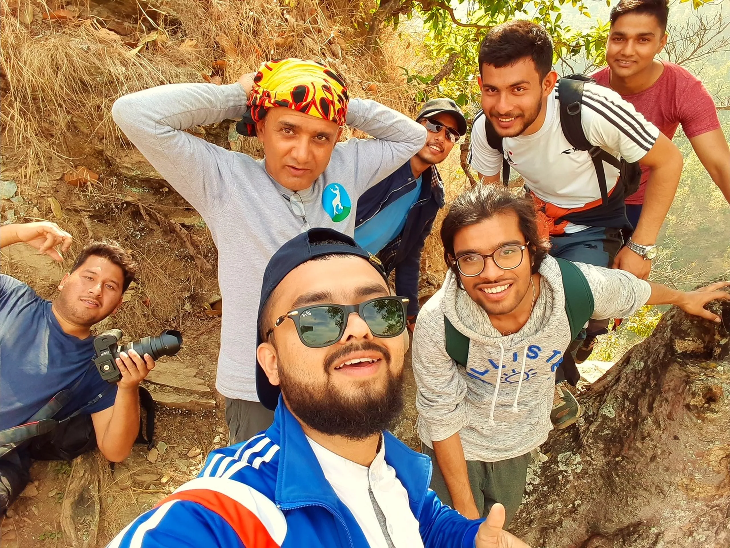 A lively, energetic group selfie taken on a rocky, sunlit hillside with a rough earthen wall or cliff face as the backdrop. The same bearded man with dark sunglasses and backwards cap in the blue jacket takes the selfie in the close foreground, grinning. Behind him, five or six other young men crowd together in various poses - one wears a striking colorful cycling or sports helmet with a bright yellow-green pattern, making his head pop visually in the cluster. Another holds a camera. The others laugh, lean in, or make expressions - one pulls a playful face. Everyone is in good spirits, faces close together, the energy of the group palpable. The rocky red-brown earth wall behind them gives the image a raw, outdoor texture. It is an unmistakably joyful group moment mid-trek.
