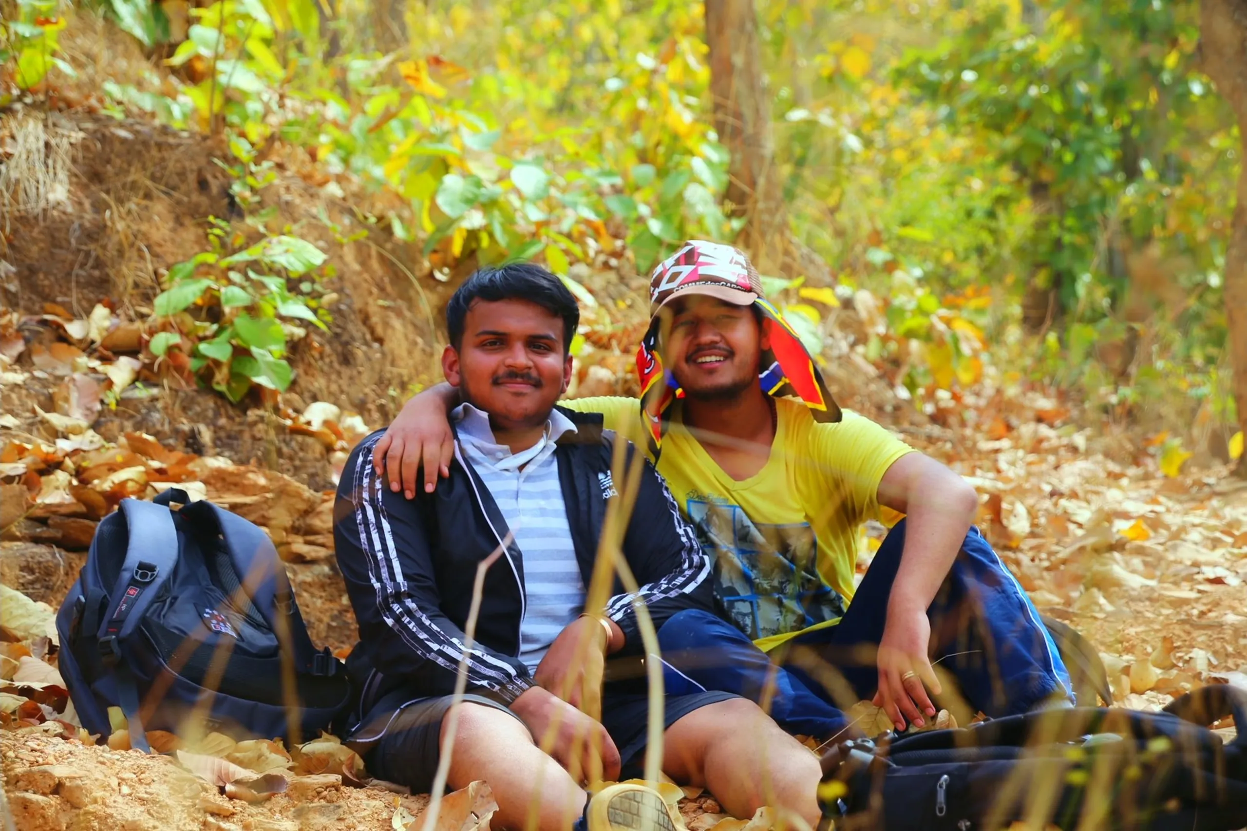 A warm, natural portrait of two young men sitting together on the dry forest floor, both facing the camera with broad, genuine smiles. The man on the left wears a dark striped or patterned collared shirt and has his arm wrapped affectionately around the shoulders of the man on the right - a gesture of easy friendship. The man on the right wears a bright yellow t-shirt and has a colorful bandana, cloth, or patterned hat tied around his head, giving him a cheerful, casual look. Backpacks and gear are visible beside them on the ground. Behind them, the forest glows with warm golden-brown dry leaves and filtered light. The image radiates comfort, ease, and the particular warmth of good company in the outdoors.