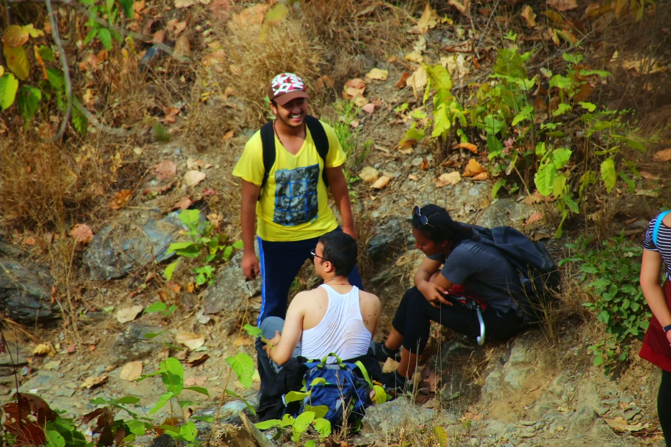 A candid action scene on a steep, rocky, dry hillside. The man in the bright yellow t-shirt and colorful sports helmet stands upright on the slope, looking down with a concerned or attentive expression at a second figure who is crouching low - possibly struggling, resting, or navigating a difficult section of the rocky terrain. A third person with a backpack is also on the slope nearby. The ground is rough and uneven - pale grey rock mixed with dry red earth and sparse dry vegetation. The interaction between the standing figure watching and the crouching one below captures a real moment of the physical challenge and mutual support of group trekking on difficult terrain.