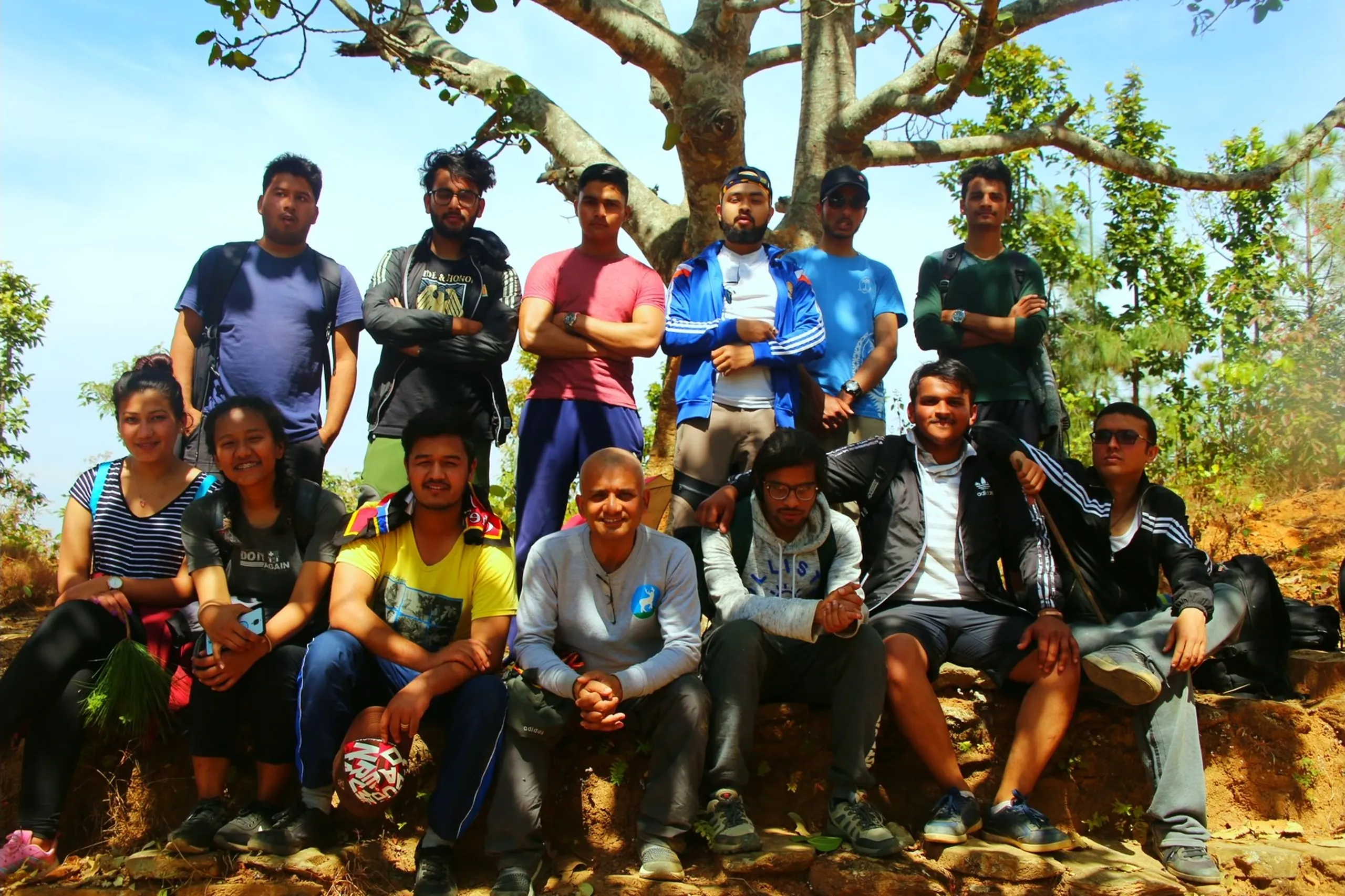 A large full group photograph of approximately thirteen to fifteen people - men and women - posed together under a wide, bare-branched tree on dry, rocky ground. The group is arranged in two informal rows: those in the back stand, those in front sit or crouch. Faces are varied and expressive - most smiling, all looking at the camera. The group includes at least two women visible in the front row. Clothing is a mix of casual trekking gear - jackets, hoodies, t-shirts in blue, yellow, black, grey. The tree above provides a natural canopy. The dry red-brown earth, scattered rocks, and open sky give this the feel of a full trekking group summit photo - the whole crew, documented together at a memorable point on the trail.