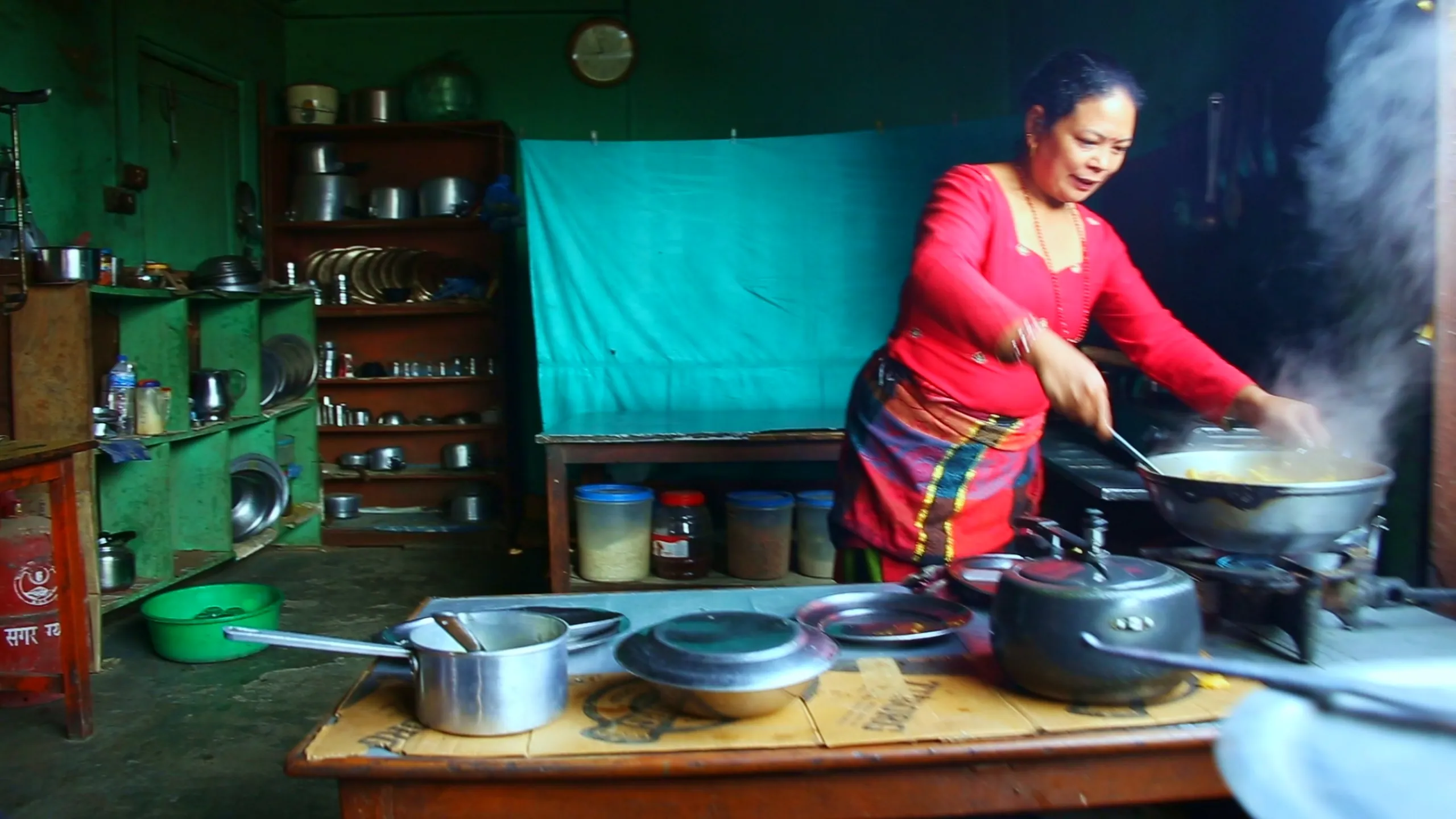 A busy, vibrant traditional Nepali kitchen in full cooking mode. A woman stands at the center, wearing a bright red full-sleeve top and a colorful patterned skirt or lungi. She is actively stirring or tending to food in large round pans or woks on a gas stove, her body slightly turned and her movements energetic. Steam or smoke rises dramatically from the cooking, filling the upper part of the image with a soft haze. The kitchen behind her is full of life - shelves lined with metal pots, pans, and utensils; a teal-green curtain separates one section from another; a round clock on the wall; stainless steel vessels of various sizes stacked or placed on surfaces. A gas cylinder is visible near the floor. The kitchen is small but functional, warm and lived-in.