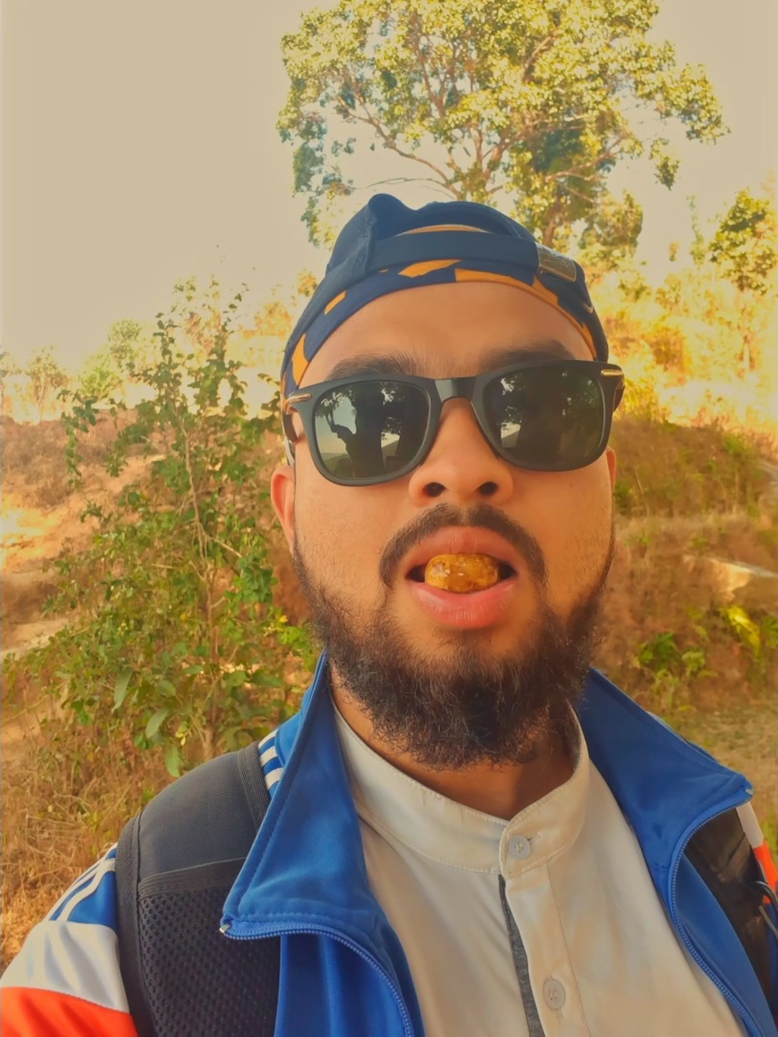 A close selfie portrait of the main bearded man of the group - dark sunglasses, backwards cap, blue jacket - in a moment of pure, unselfconscious fun. His mouth is open, and a small wild fruit is visibly placed on or between his lips or front teeth - he is in the act of tasting or eating one of the foraged fruits found in the previous images, making a slightly exaggerated or playful face at the camera. The background behind him shows a dry hillside landscape of golden-brown and pale trees and shrubs, slightly overexposed in warm light. The image is candid and funny - a man eating a wild fruit on a mountain trail, documenting the moment for posterity with a selfie.