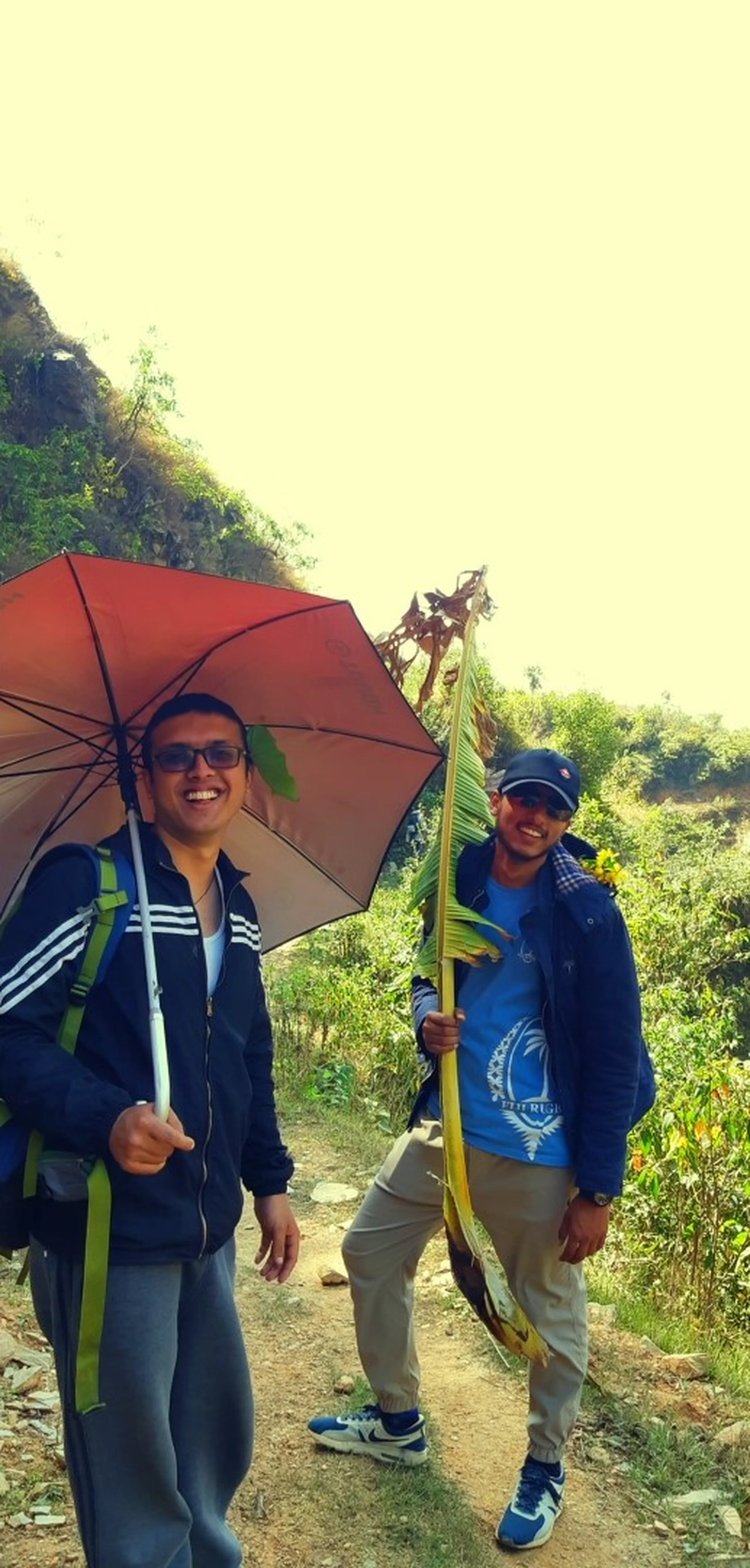 Two men walk along a bright, sunny mountain trail in a moment of cheerful absurdity. The man in the foreground - wearing a dark Adidas track jacket, grey trousers, and sunglasses - carries an open red umbrella above his head against the sun, grinning widely and looking at the camera. Behind him, the second man in a blue jacket carries an enormous fresh green banana leaf or palm frond over one shoulder like a flag or walking staff - its long, wide, vivid green blade extending well above his head. Both men look delighted with themselves. The background is a bright, slightly overexposed rocky hillside with green vegetation. The image is full of playful energy - two friends making the trail their own.
