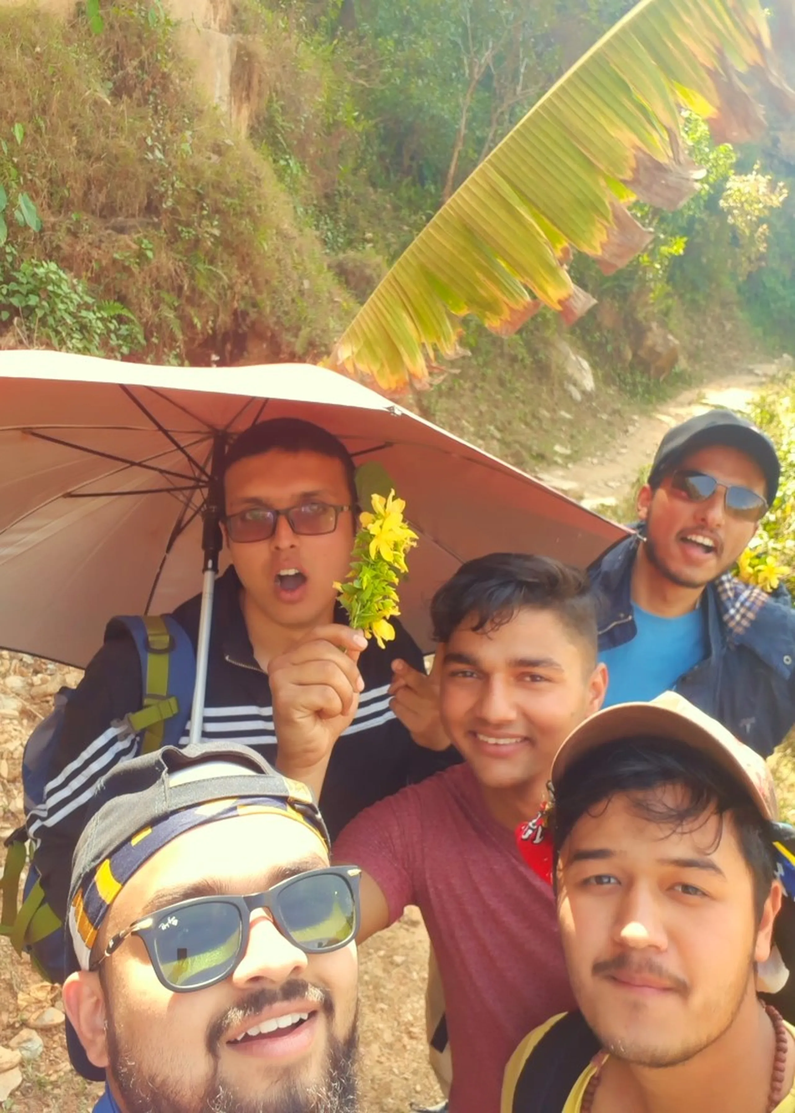 A chaotic, joyful close group selfie of four young men on a rocky hillside trail, all crowded into the frame at once. The man taking the selfie - dark sunglasses, colorful patterned cap - is in the sharp foreground, mouth open in laughter. Behind him, three faces cluster close: one man in glasses and a red t-shirt grins broadly; another holds a small bright yellow wildflower up near his chin with theatrical flair; a fourth wears a cap and sunglasses and smiles from the side. Above their heads, the red umbrella is visible, and the edge of the large green banana leaf/frond appears behind them. A rocky hillside and a fragment of banana tree leaf make up the background. The image is loud, expressive, and full of genuine, unscripted happiness.